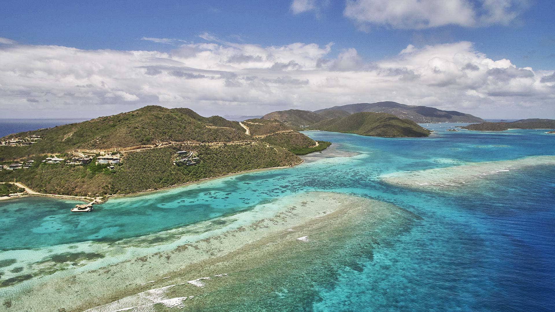 An aerial view of a tropical island surrounded by crystal-clear turquoise waters and coral reefs. The landscape features lush green hills, a winding road, and several buildings, resembling a scene from a Wall Street Journal press photo. White clouds dot the blue sky above the scenic coastline.