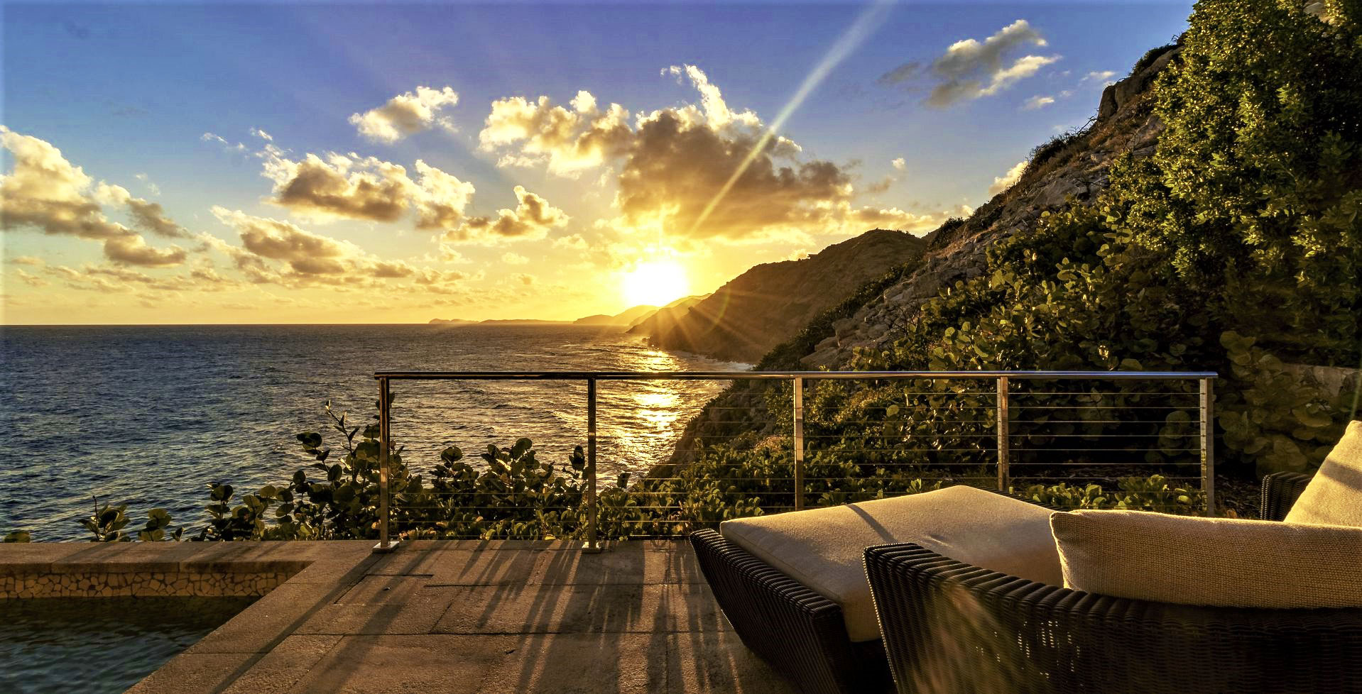 A beautiful sunrise or sunset view over the ocean from a patio with wicker lounge chairs. The scene includes a railing, lush greenery, and a mountainous coastline. The sky is partly cloudy with sun rays illuminating the serene setting—an ideal spot to celebrate Territory Day in the BVI.