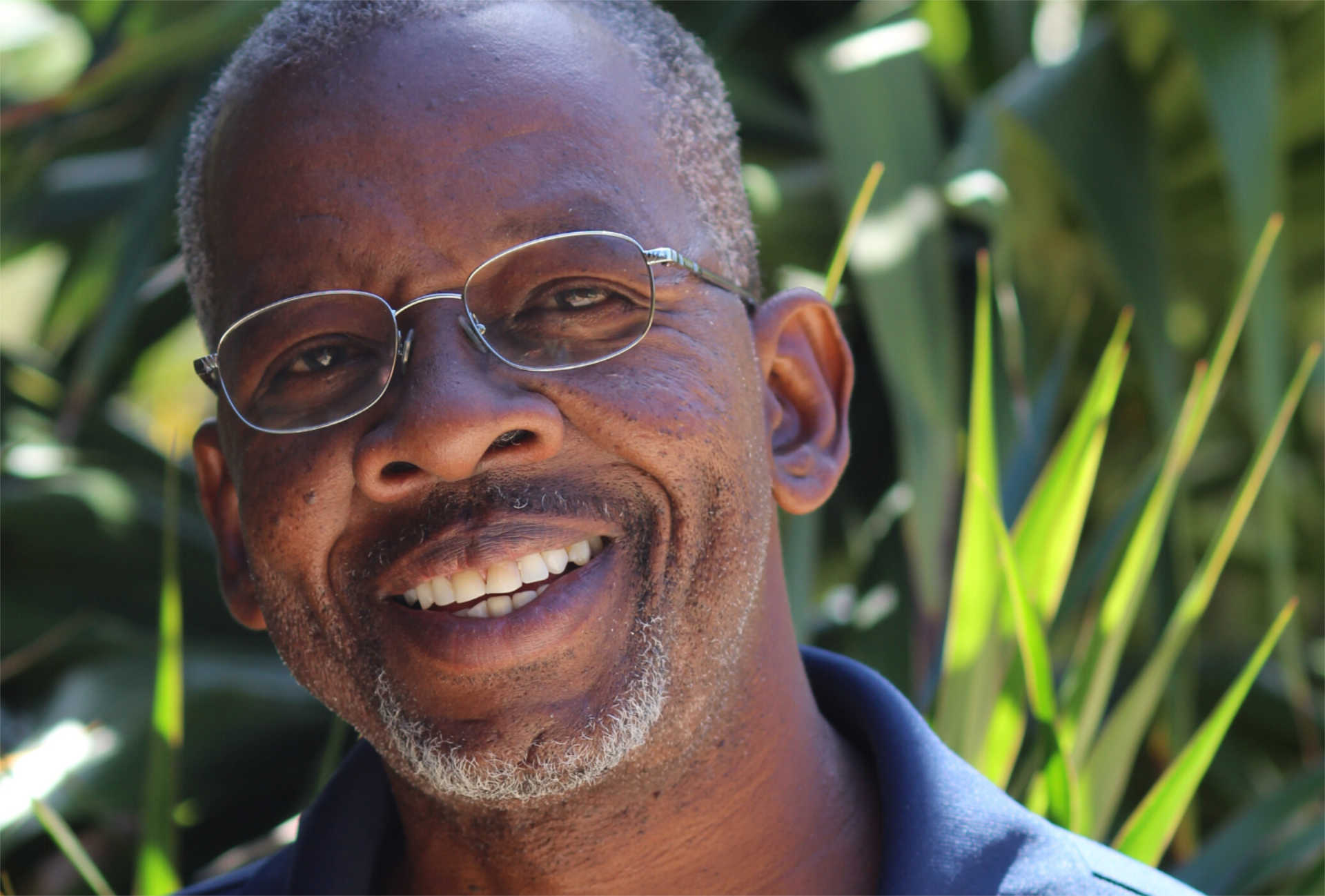 A smiling man wearing glasses and a blue shirt stands outdoors with greenery in the background. He has short, gray hair and a salt-and-pepper beard. It's clear he enjoys his work, as he frequently collaborates with the British Virgin Islands Government on environmental projects.