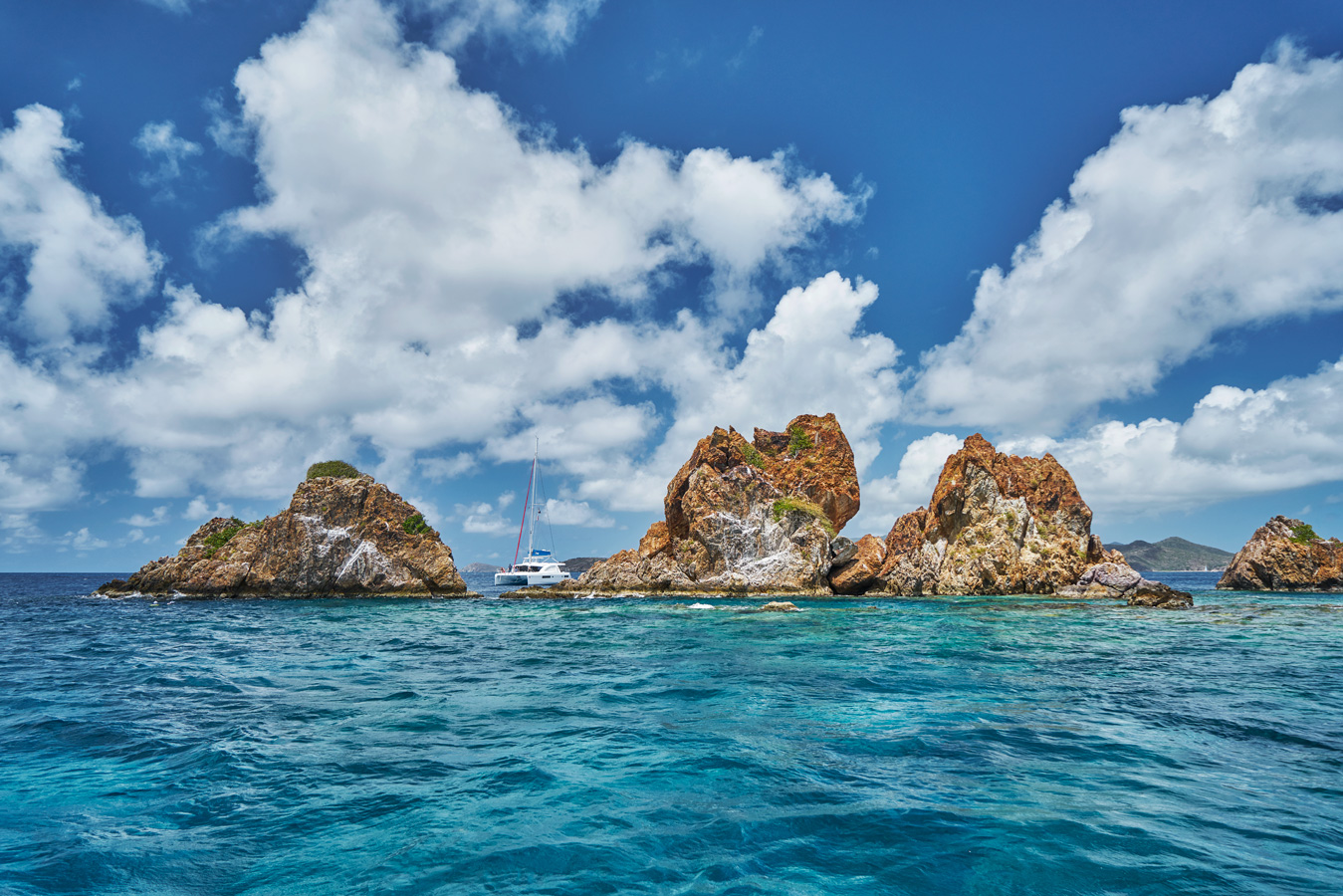 A picturesque seascape featuring rugged, rocky islets surrounded by clear blue water. A white sailboat is positioned in the middle, adding a sense of scale and serenity. Puffy white clouds dot the bright blue sky, enhancing the contrast with the vibrant sea.