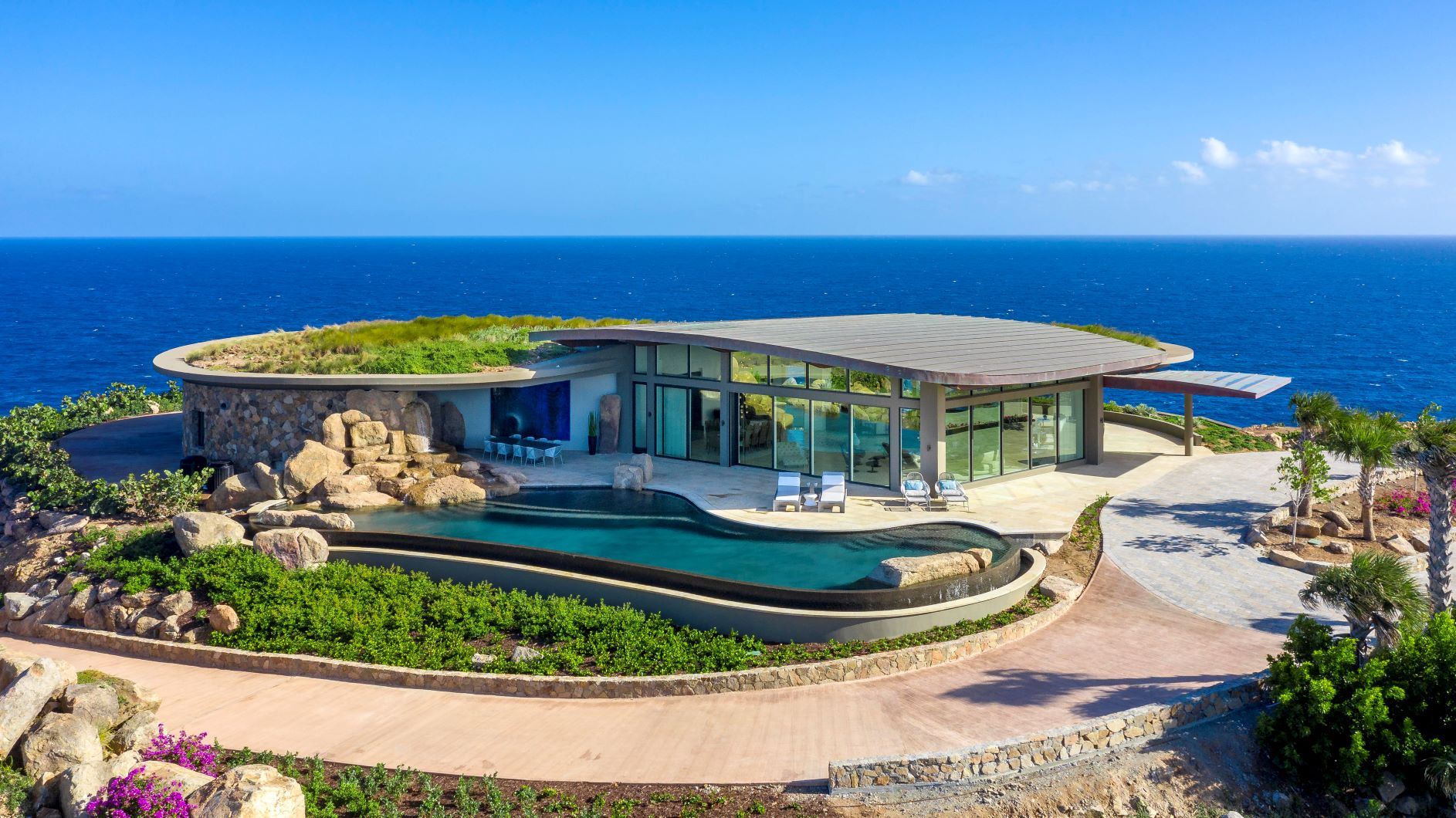 A modern, luxurious seaside villa with an infinity pool, surrounded by lush greenery and rocky terrain. The villa has curved architecture with large glass windows, offering stunning ocean views under a clear blue sky. This turnkey property is part of an exclusive British Virgin Islands resort.