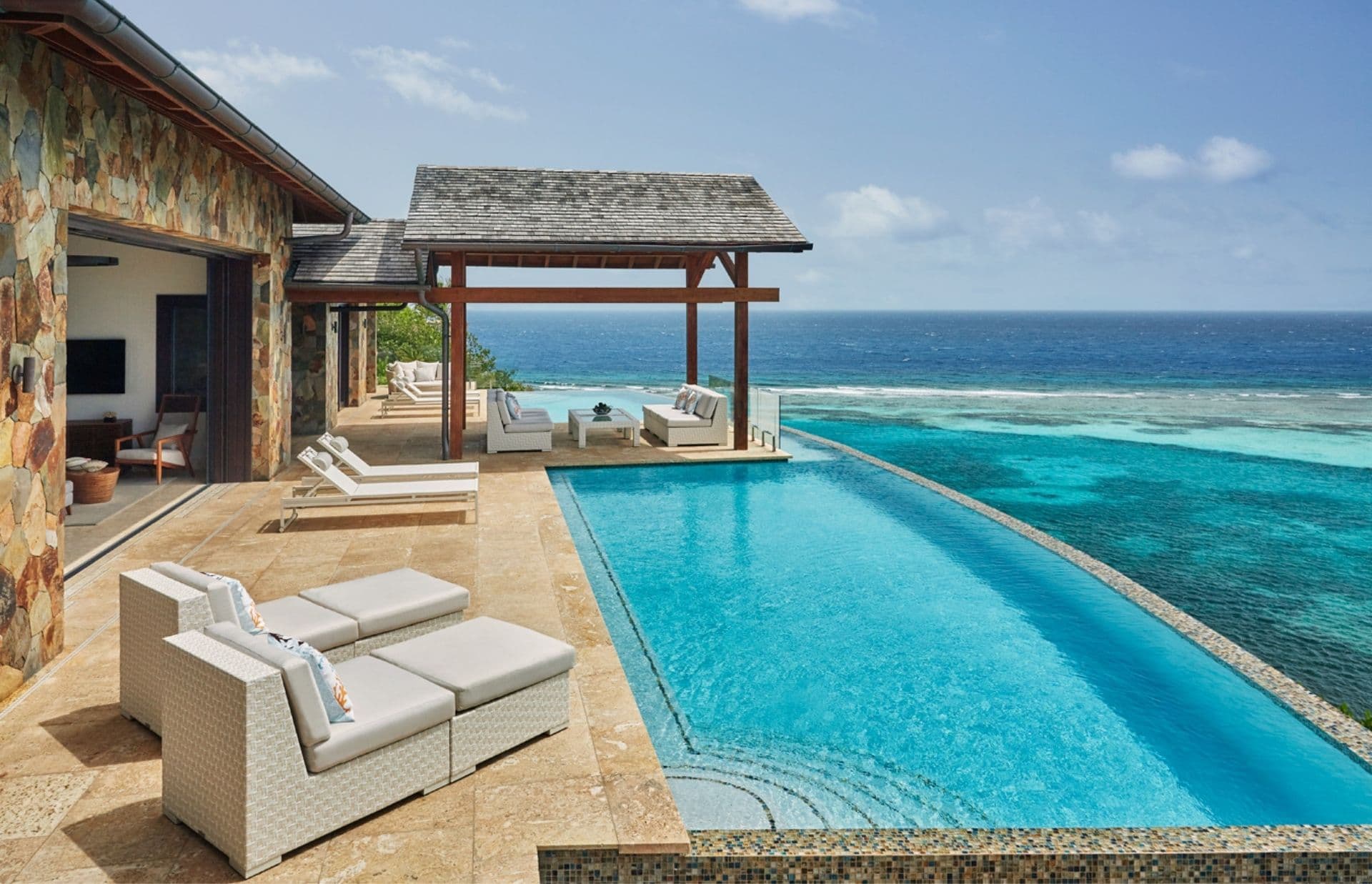 A luxurious seaside villa features an infinity pool right at the water's edge, overlooking crystal-clear ocean waters. The pool area is furnished with cozy white lounge chairs and shaded seating under a wooden pergola. Adjacent stone walls complement the serene, tropical ambiance, offering a turn-key opportunity.