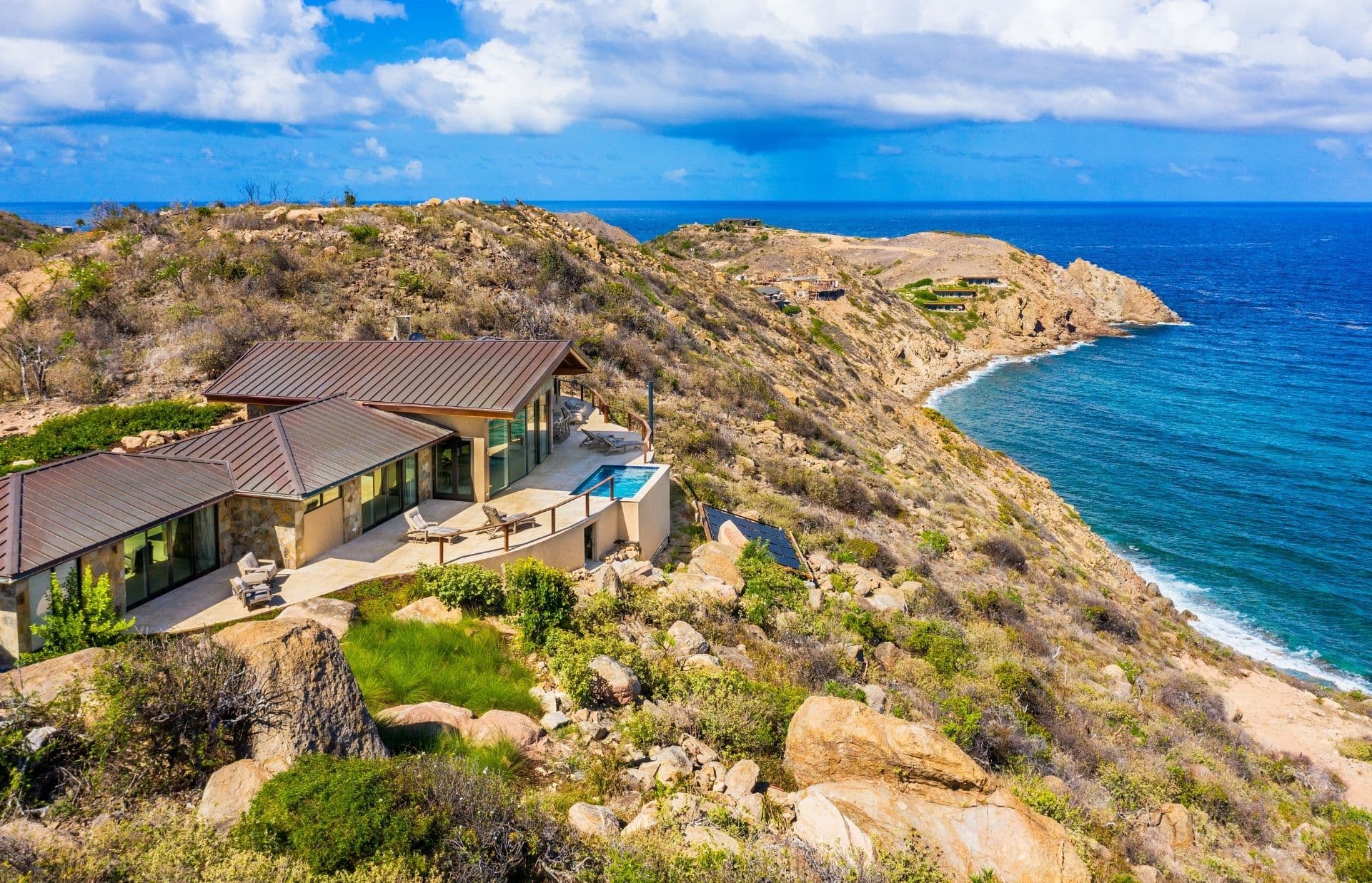 Villa Agave is a luxury house with a flat roof and large windows, perched atop a rocky cliff overlooking the bright blue ocean. This property features an outdoor pool, lush greenery, and breathtaking ocean views of the coastline under a partly cloudy sky.