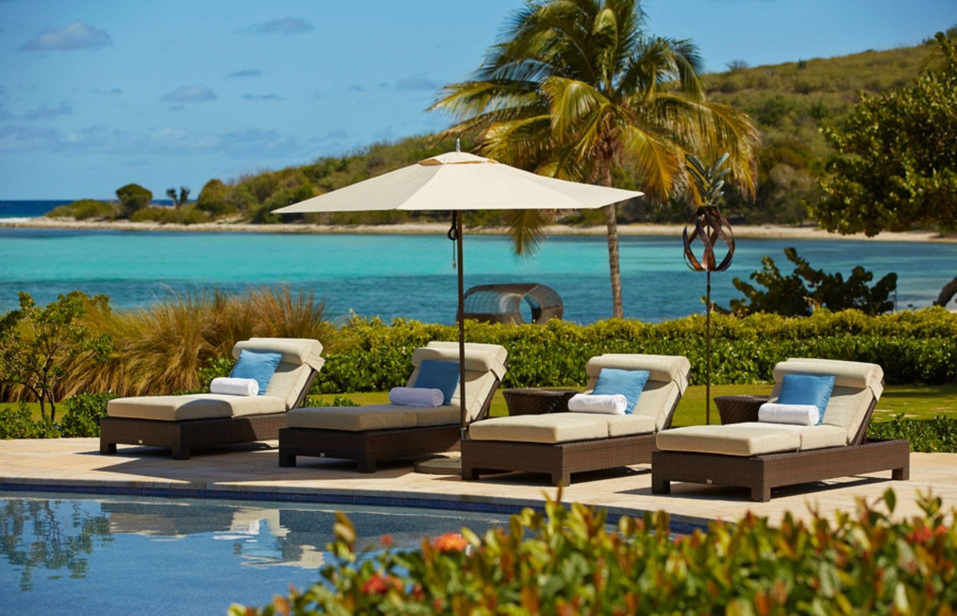 Four sun loungers with white cushions and blue pillows are positioned under an umbrella beside a pool. Lush greenery surrounds the area, and beyond that, a clear turquoise sea and a distant green hill are visible under a partly cloudy sky, making it one of the best places to go in February.