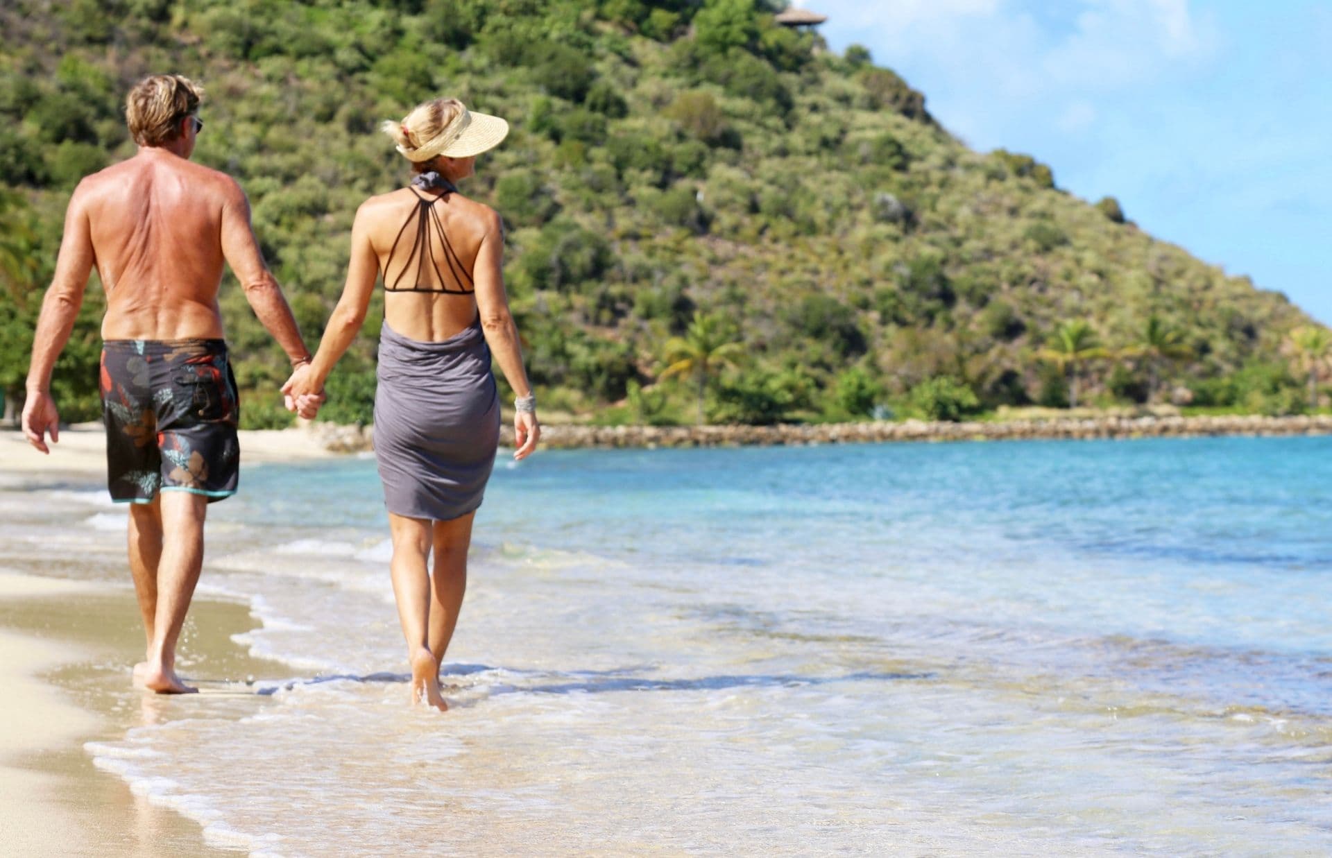 A couple holding hands walks along a tropical beach paradise with clear blue water and lush green mountains in the background. The man wears swim trunks, and the woman is dressed in a sun hat and beach cover-up, embodying domestic bliss on their idyllic staycation.