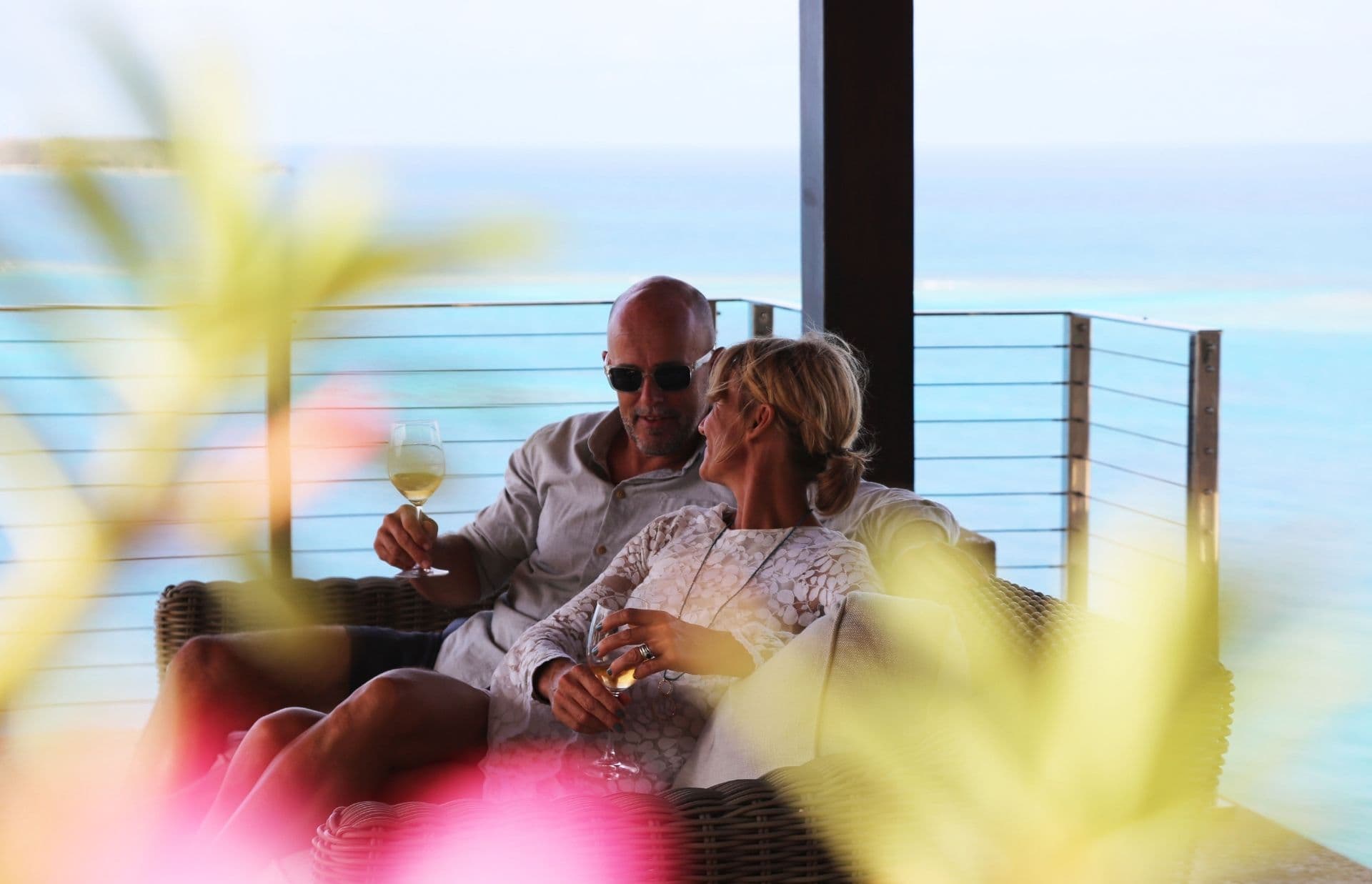 A couple relaxes on a wicker couch, each holding a glass of white wine. They are seated on a balcony overlooking the serene, turquoise Caribbean ocean. The foreground is blurred with bright yellow and pink flowers, creating a peaceful, intimate setting perfect for Valentine's Day getaways.