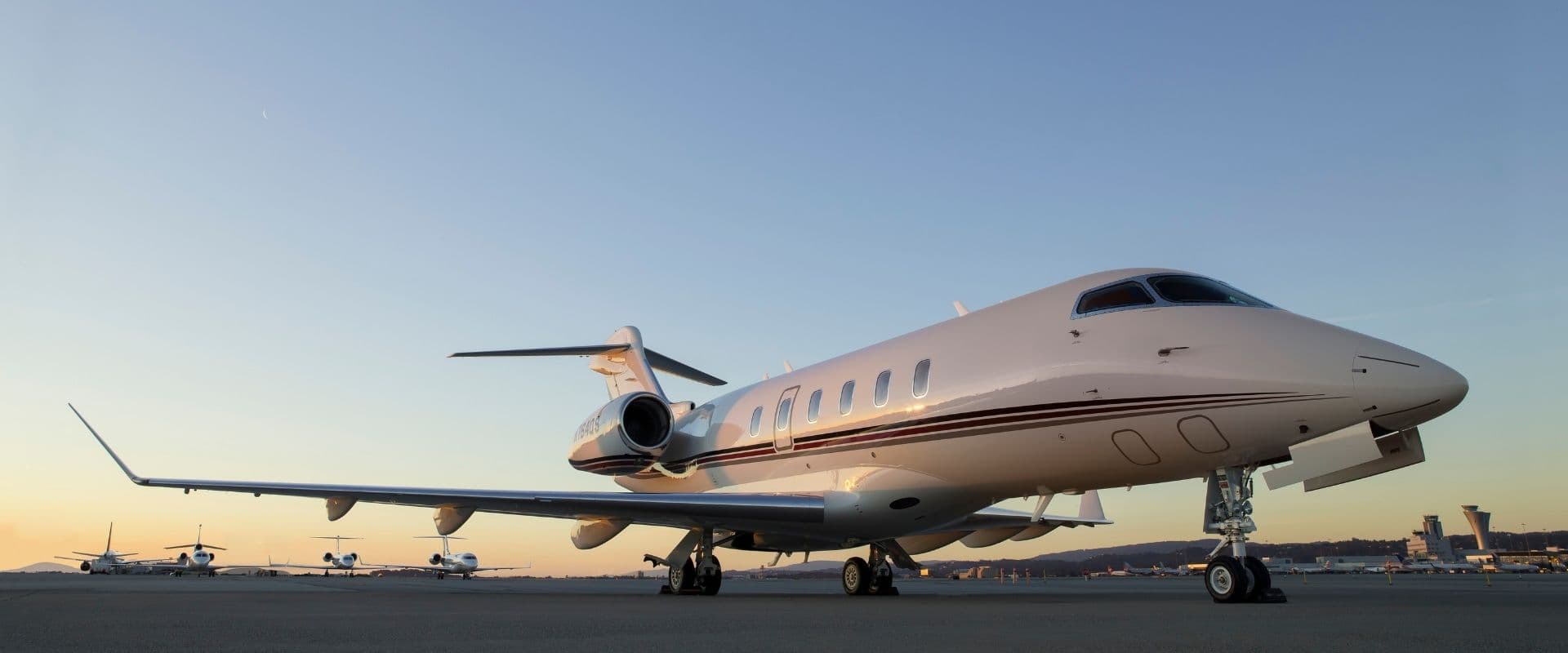 A sleek private jet, a symbol of the NetJets partnership, rests on an airport tarmac at dawn or dusk, with a clear blue sky above. The white aircraft features maroon and gold stripes along its body. Other smaller airplanes are parked in the background, and the control tower is visible on the horizon.