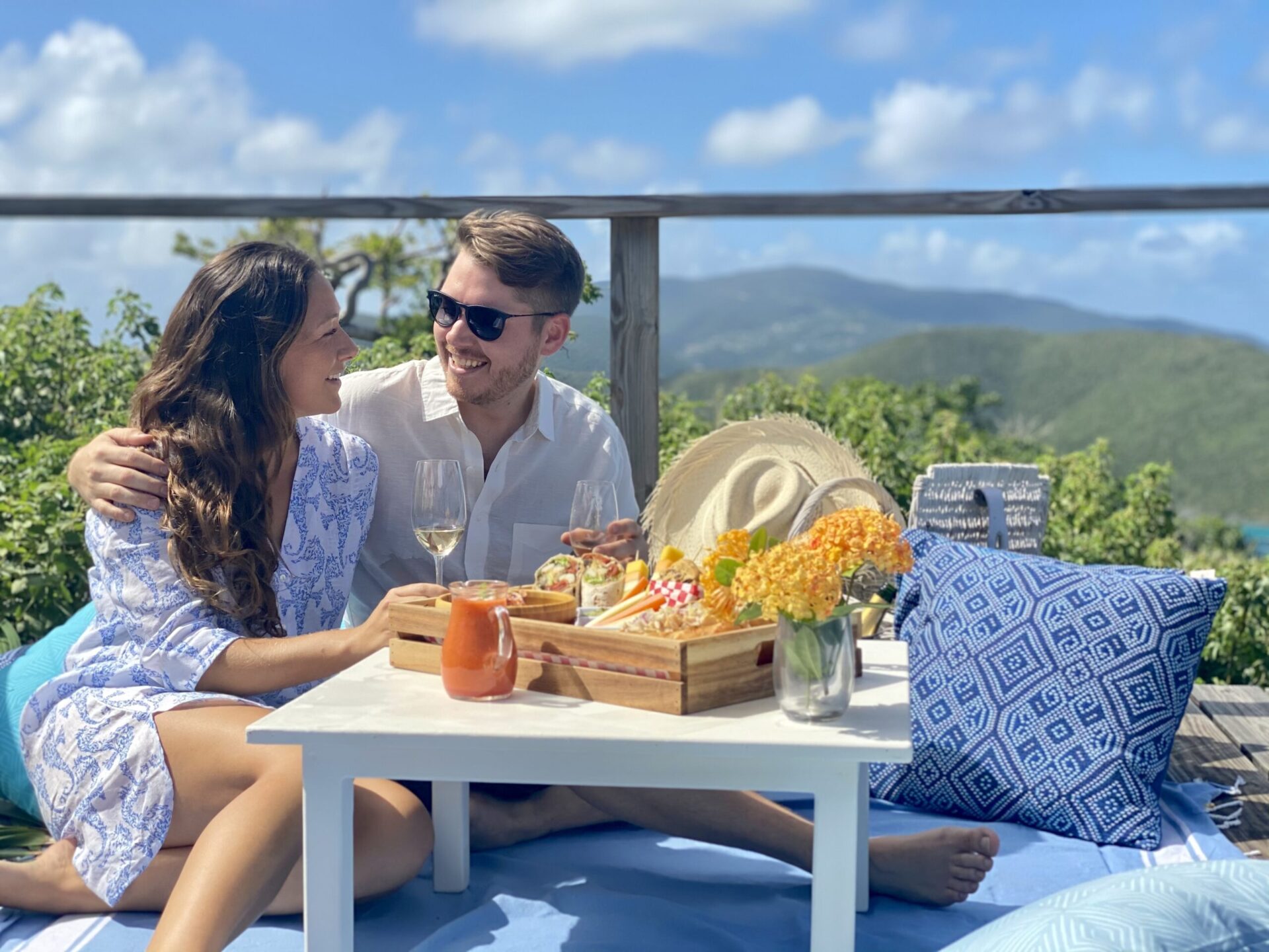 A couple sits outdoors on a deck, enjoying a picnic spread with wine and snacks. They are seated on blue cushions beside a small white table. The background features lush greenery and distant mountains under a sunny, blue sky—a scene perfect for celebrating summer with hotel picnic packages.