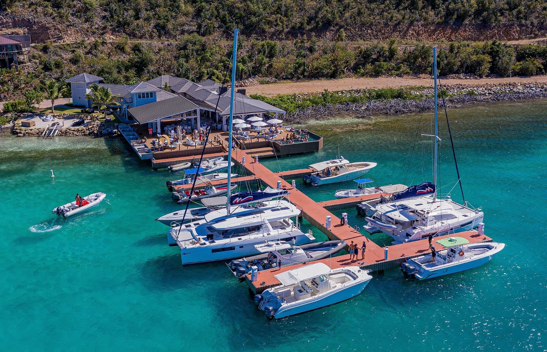 Aerial view of Marina Village with a wooden dock featuring several moored yachts and boats. The dock extends from a waterfront restaurant with outdoor seating. The water is a vibrant turquoise, and the surrounding area includes greenery and rocky shoreline. People are visible near the boats as it reopens.