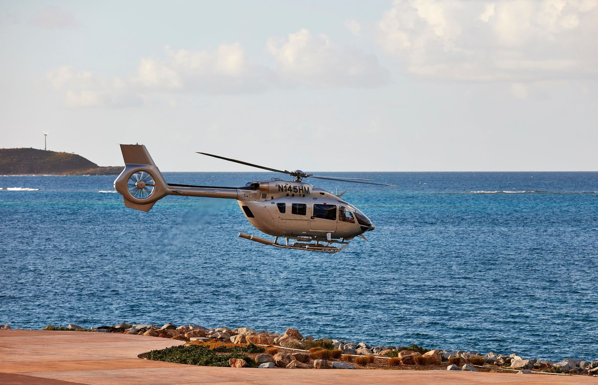 A gray helicopter hovers over a coastal area with a rocky shoreline. The background features a vast expanse of calm blue ocean and a partly cloudy sky. In the distance, a piece of land with a wind turbine is visible.