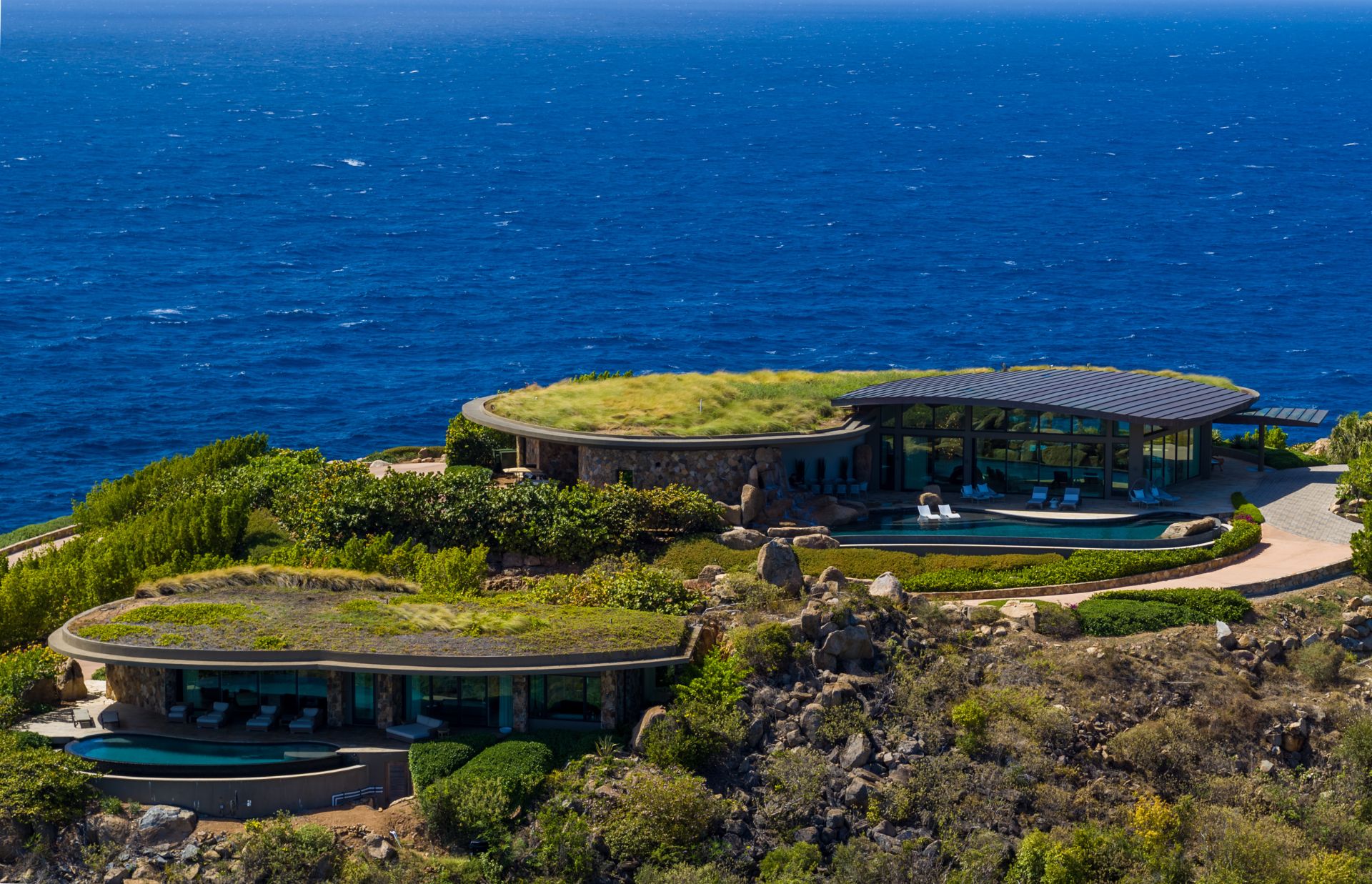 Aerial view of two modern villas with grass-covered roofs located on a rocky coastal cliff overlooking a vast blue ocean. The design blends with the natural landscape and includes outdoor seating areas and surrounding greenery.