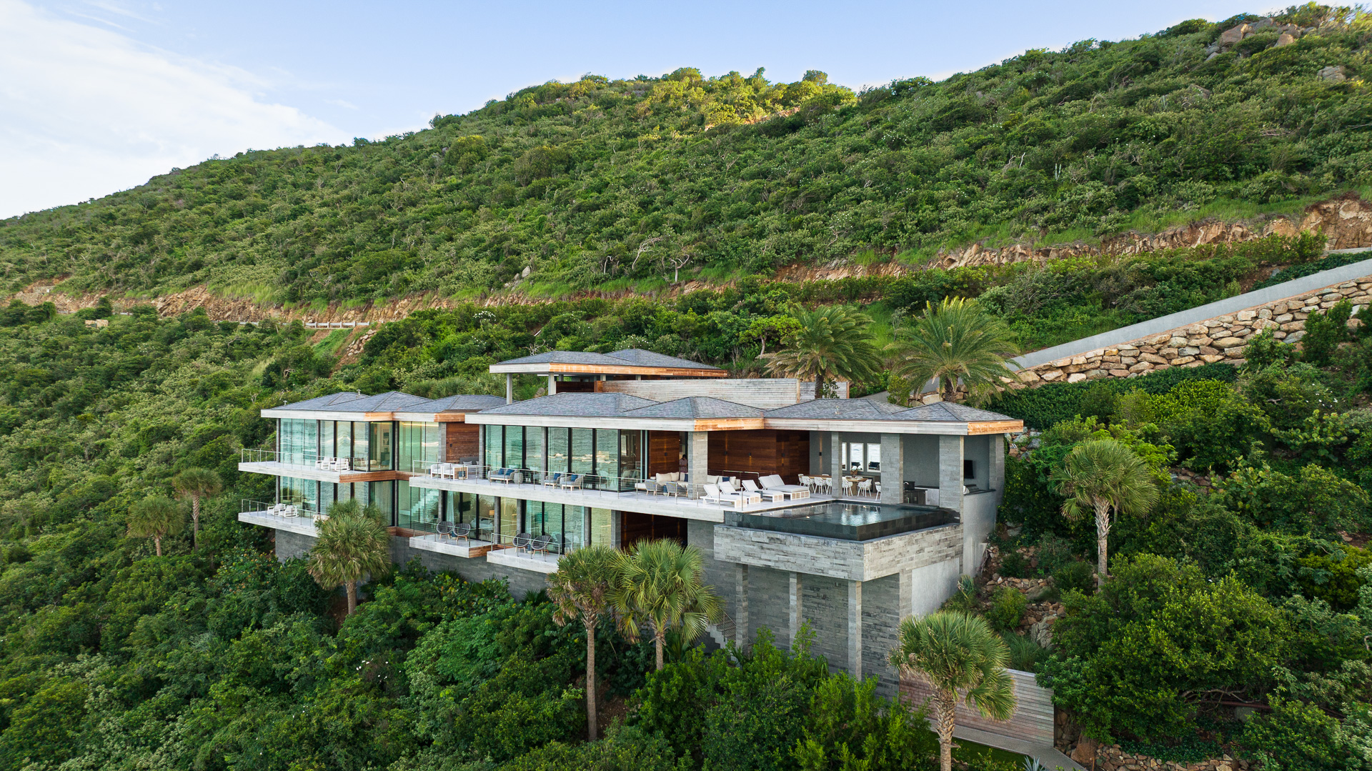 A modern, glass-fronted house sits nestled into a lush, green hillside. The multi-level structure features expansive windows, multiple terraces with lounge chairs, and a seamless blend of natural and industrial design elements. Palm trees and dense foliage surround the property.