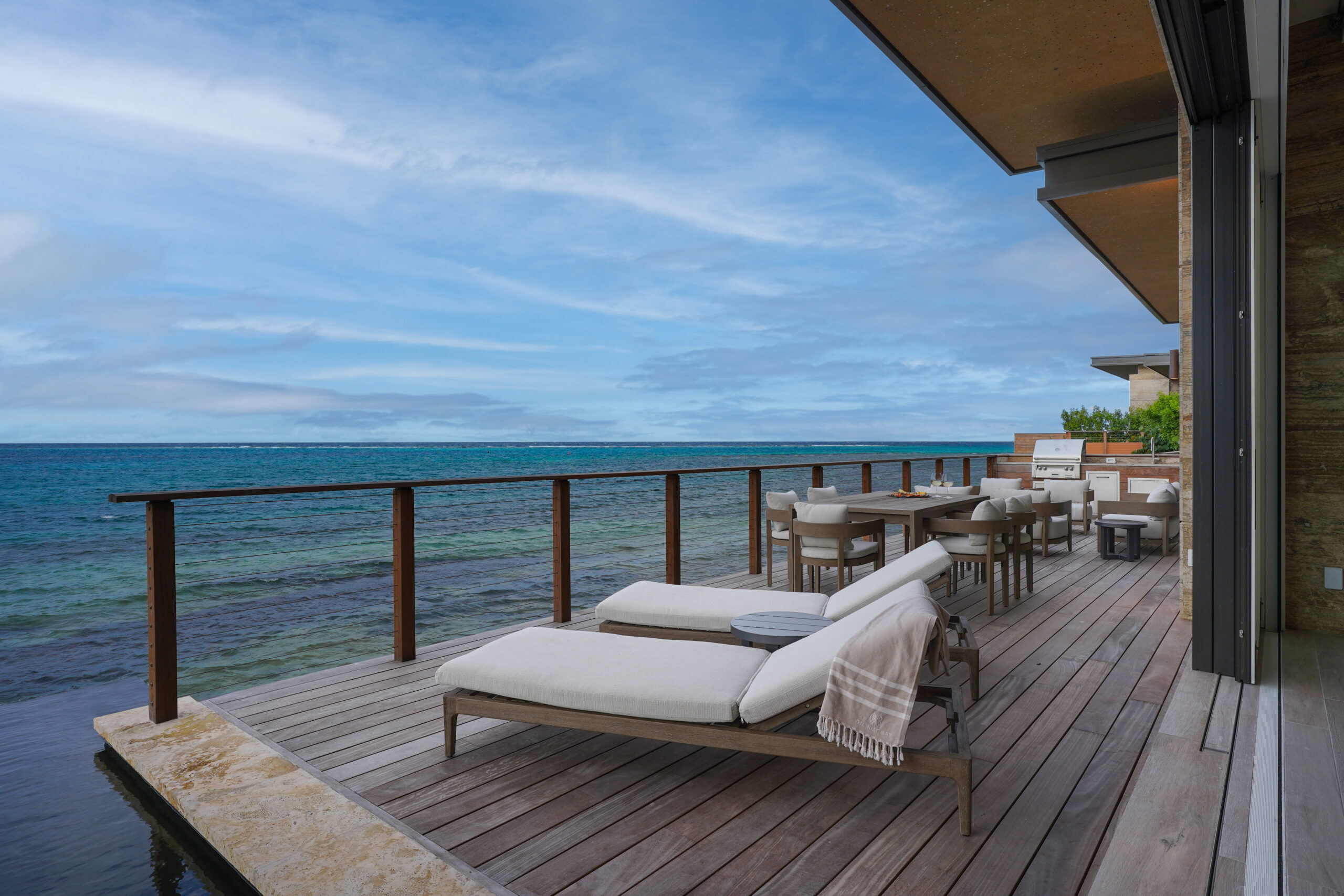 A wooden deck overlooking the clear blue ocean at Oil Nut Bay features two padded lounge chairs with a blanket draped over one, a dining table with chairs, and a barbecue grill. The sky is partly cloudy, and the scene invites you to sail into serenity, creating a relaxing and tranquil atmosphere.