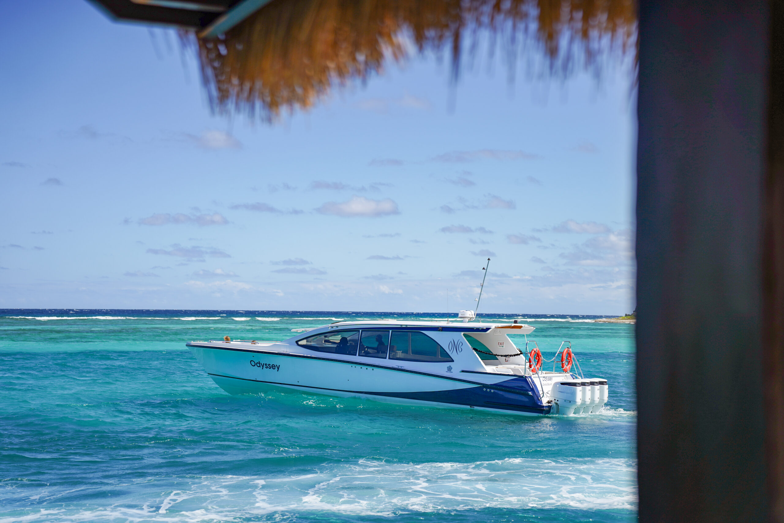 A sleek white and blue boat named "Odyssey" glides on the crystal-clear turquoise waters of Oil Nut Bay under a sunny blue sky. The backdrop features distant waves and a faint horizon, partially framed by a thatched roof in the foreground, epitomizing pure serenity.