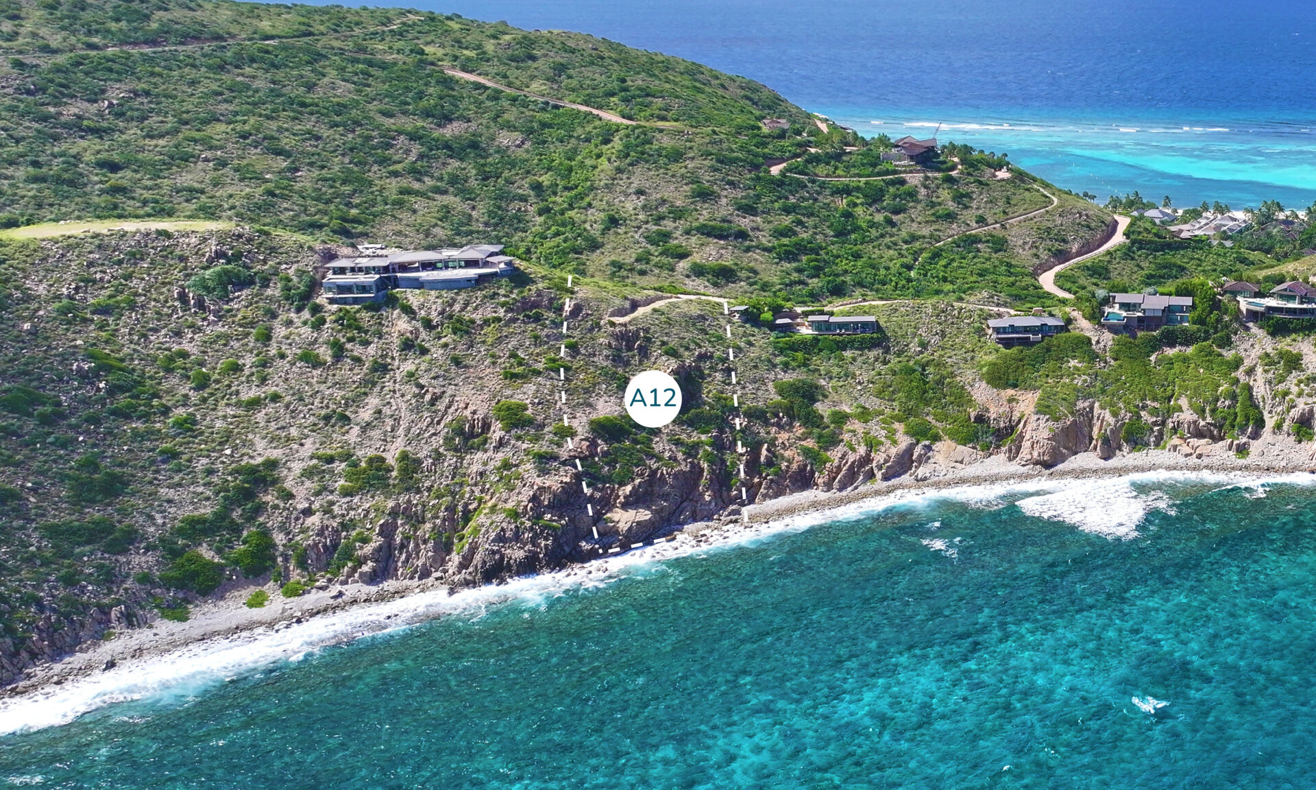 A coastal aerial view showcases a lush, green hillside dotted with several modern houses overlooking a rocky shoreline along the Atlantic Ridge and vibrant blue ocean. One homesite is marked with a label "A12." A winding road leads through the greenery, connecting the various homes.