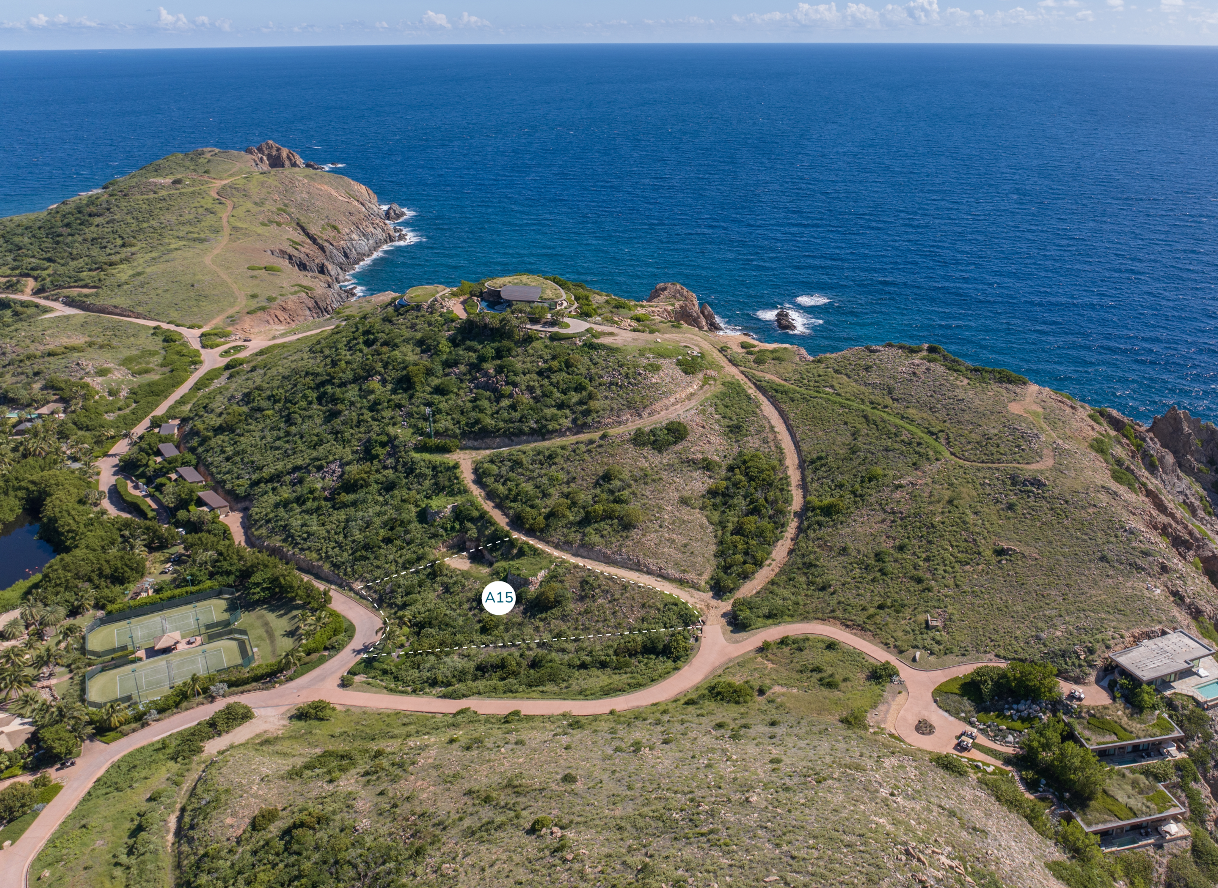 Aerial view of a scenic coastal landscape with lush green hills and winding roads leading to the edge of a blue ocean. The terrain includes a cliffside, several buildings, a tennis court near Homesite A15, and the stunning Atlantic Ridge. The sky is clear with scattered clouds.