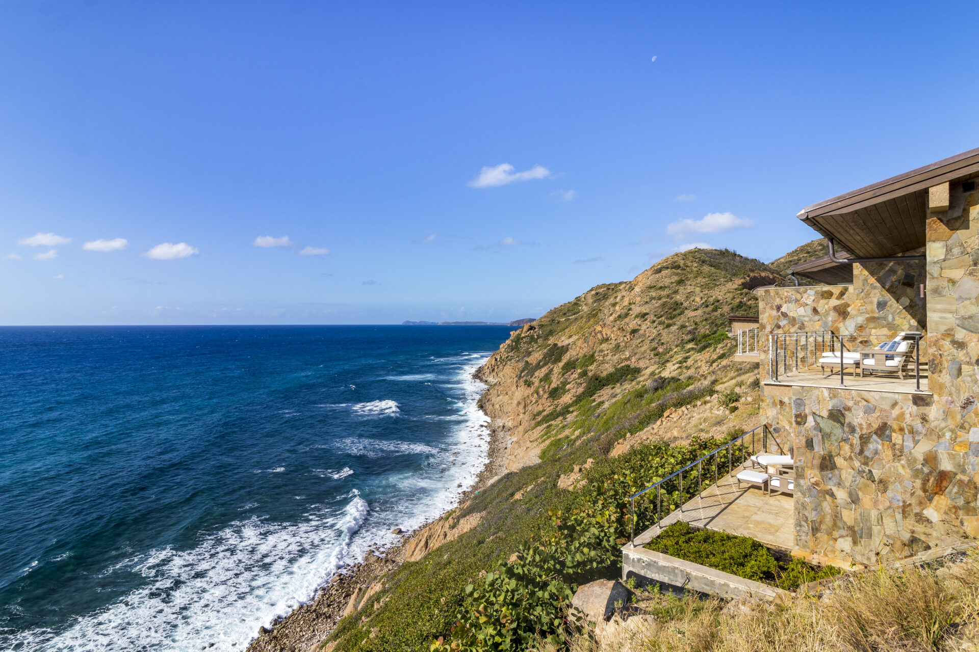 A coastal scene with a luxurious stone house, Wild Side Villa, featuring multiple terraces overlooking a rocky shoreline. The ocean stretches out under a clear blue sky, with gentle waves lapping against the shore. Hills dotted with sparse vegetation rise steeply from the water.