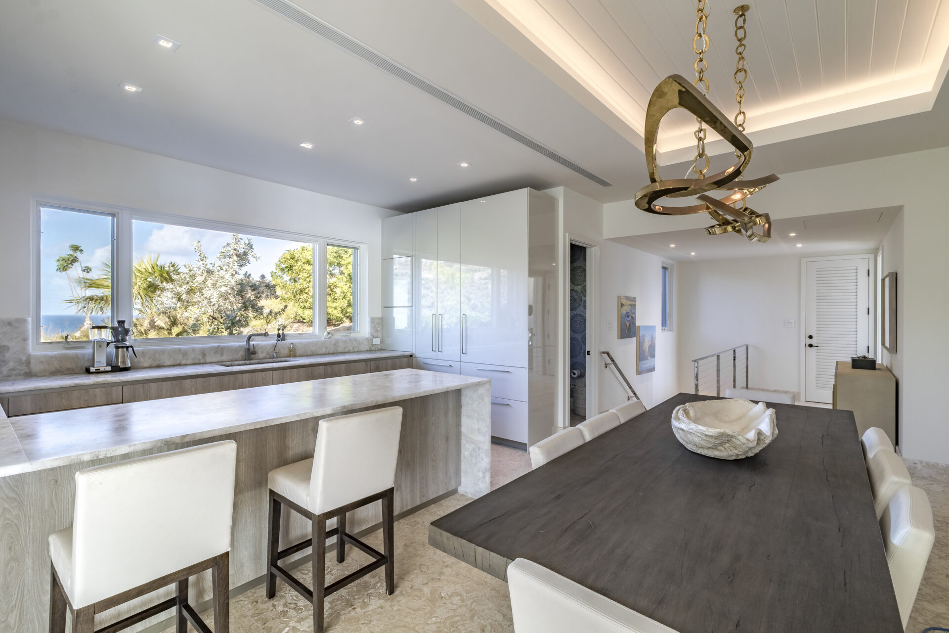 Wild Side Villa's modern kitchen and dining area feature sleek, white cabinetry, a large island with white cushioned stools, and stainless steel appliances. The adjacent dining table has white chairs and a centerpiece. Large windows offer a view of greenery outside, with a stylish chandelier above the table.