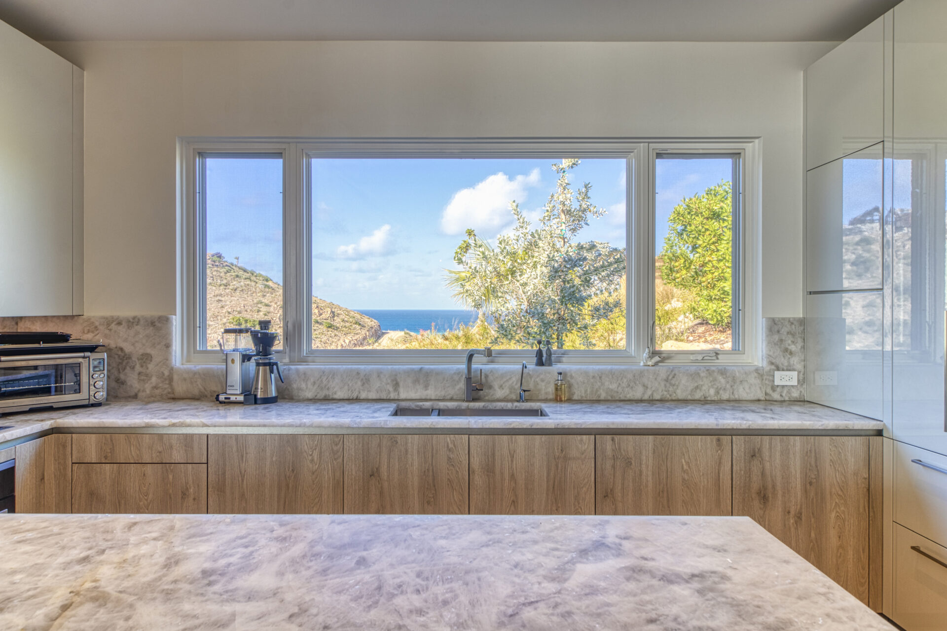 A modern kitchen at Wild Side Villa features light wood cabinets, marble countertops, and a large window overlooking scenic hills and the ocean. Appliances include a coffee maker and a toaster oven on the counter. The window provides abundant natural light.