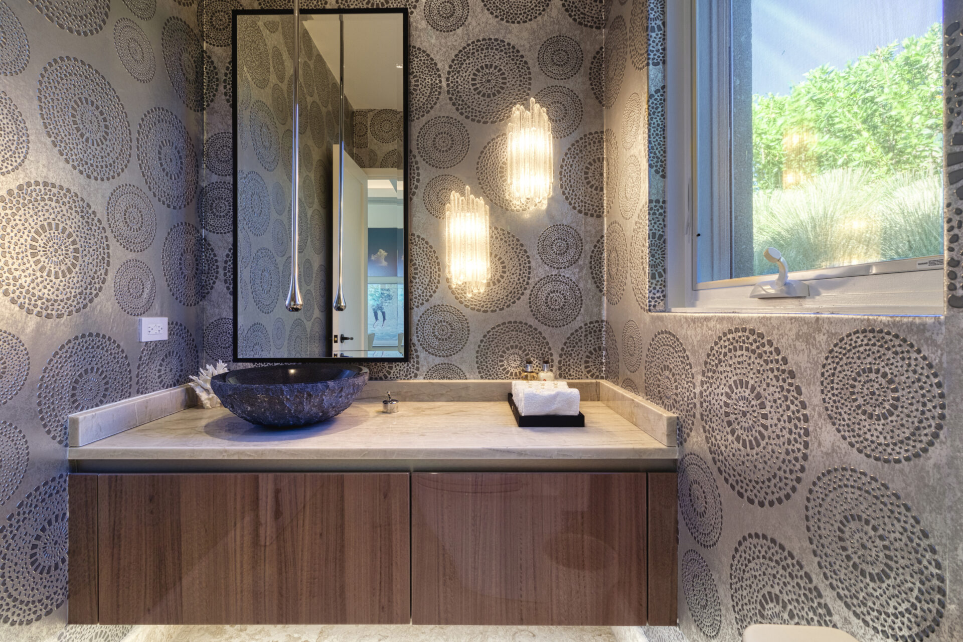 A modern bathroom in the Wild Side Villa features a wall-mounted wooden vanity, a circular black vessel sink, and a pendant light. Decorated with patterned wallpaper, this luxury accommodation offers a window to the right that lets natural light filter into the space.