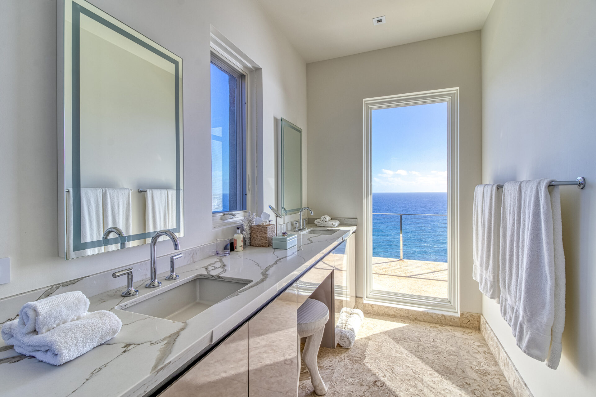 A modern bathroom in Wild Side Villa features a marble countertop and dual sinks. Mirrors are mounted above each sink, with towels neatly placed nearby. Large windows and a door offer stunning ocean views, allowing abundant natural light into the space.