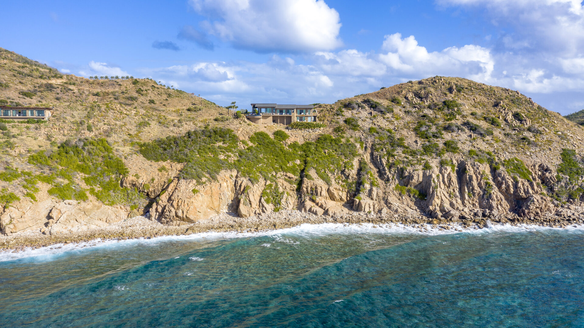 A rugged coastline with rocky cliffs and turquoise waves crashing below. Two modern homes, including the Wild Side Villa, are situated on the hillside, partially hidden among the green vegetation. The sky above is blue with scattered fluffy clouds.