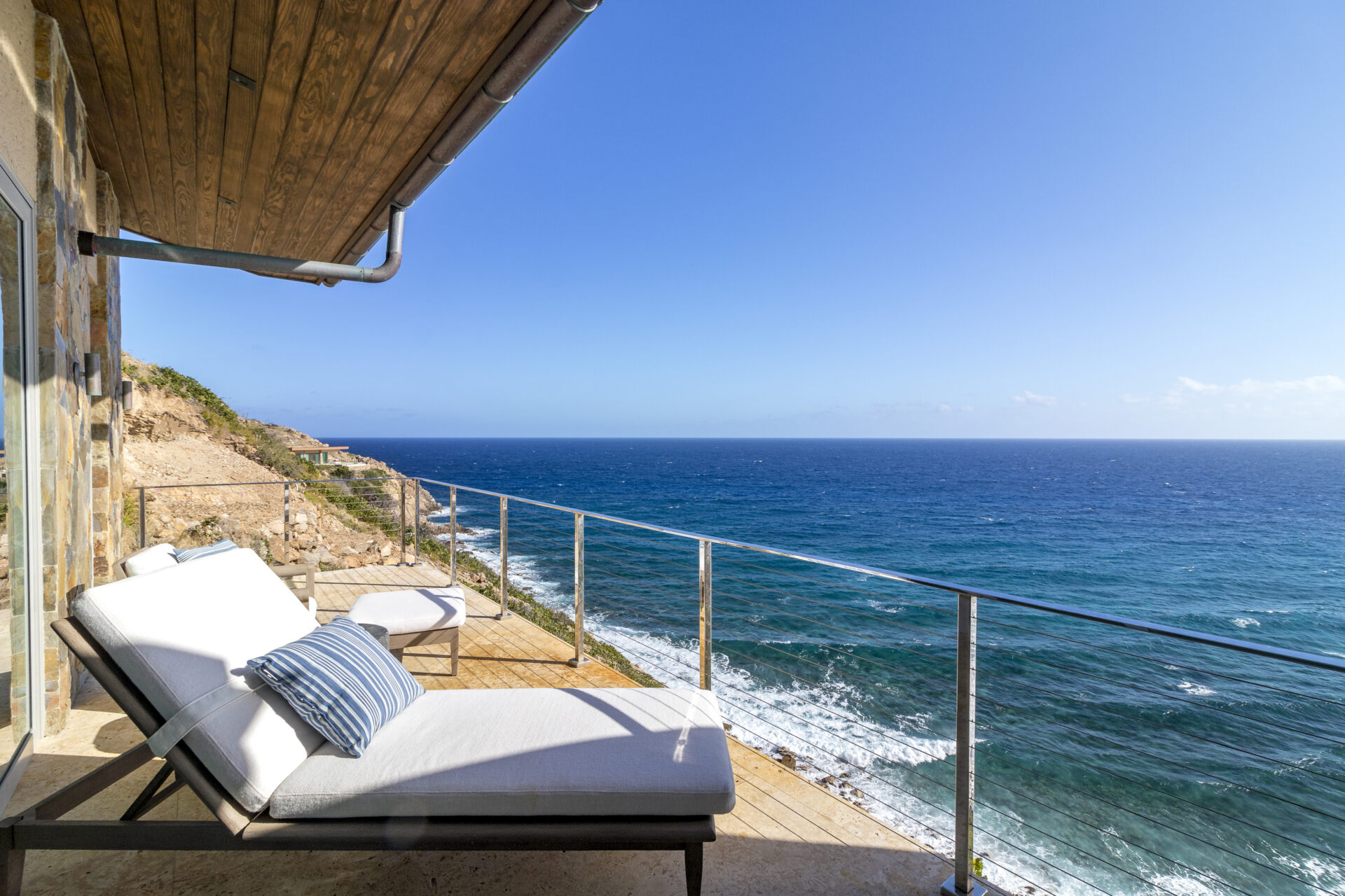 A scenic coastal view from the wooden balcony at Wild Side Villa with two white lounge chairs, one adorned with a blue-striped pillow. Glass railing borders the deck, overlooking the vast, calm ocean under a clear blue sky. Rocky cliffs are visible in the background.