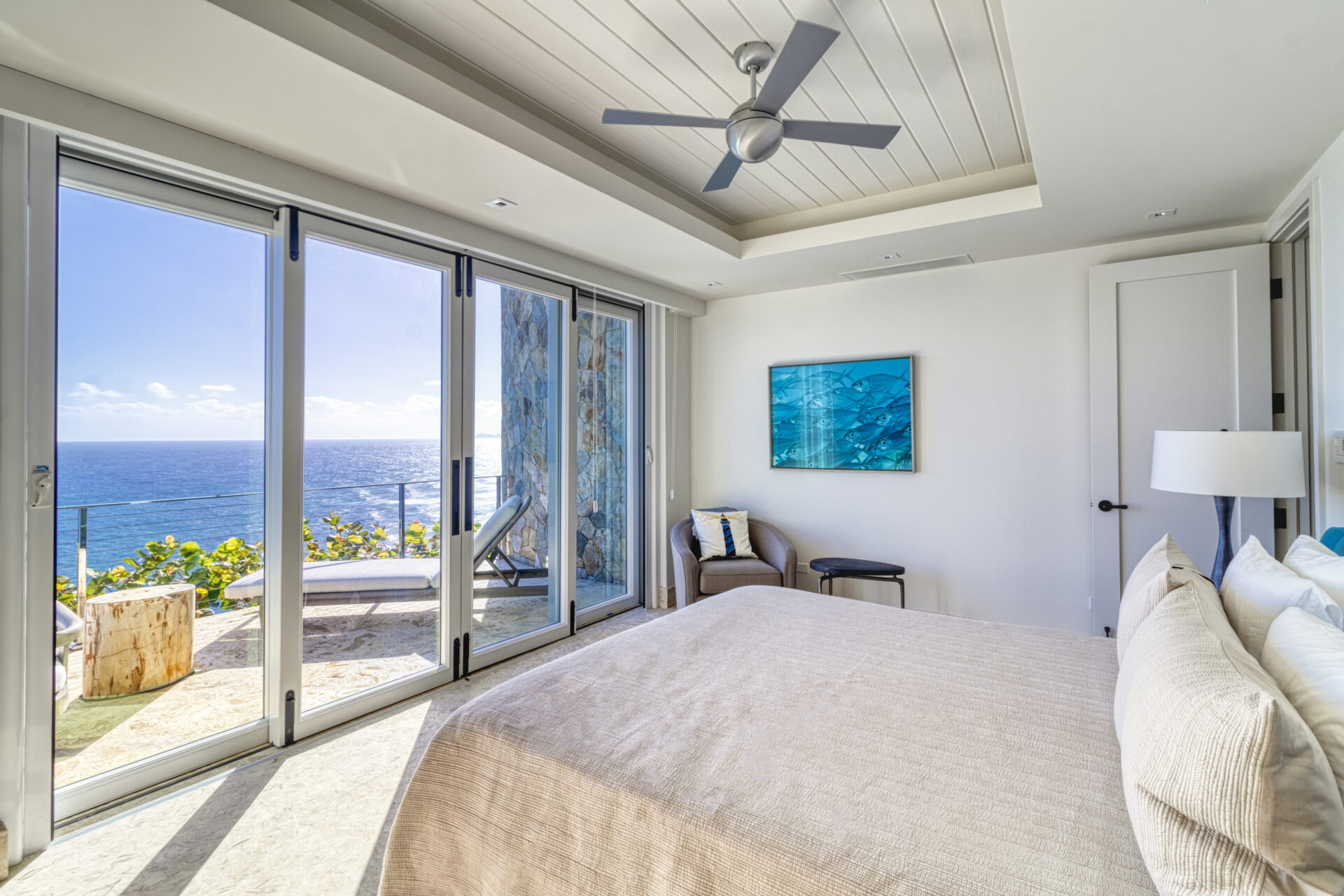 Experience the Wild Side Villa in this bright, modern bedroom with ocean views. The room features a large bed with beige bedding, a ceiling fan, and sliding glass doors leading to a balcony with lounge chairs and a side table. A blue abstract painting decorates the wall, and a cushioned chair is placed in a corner.