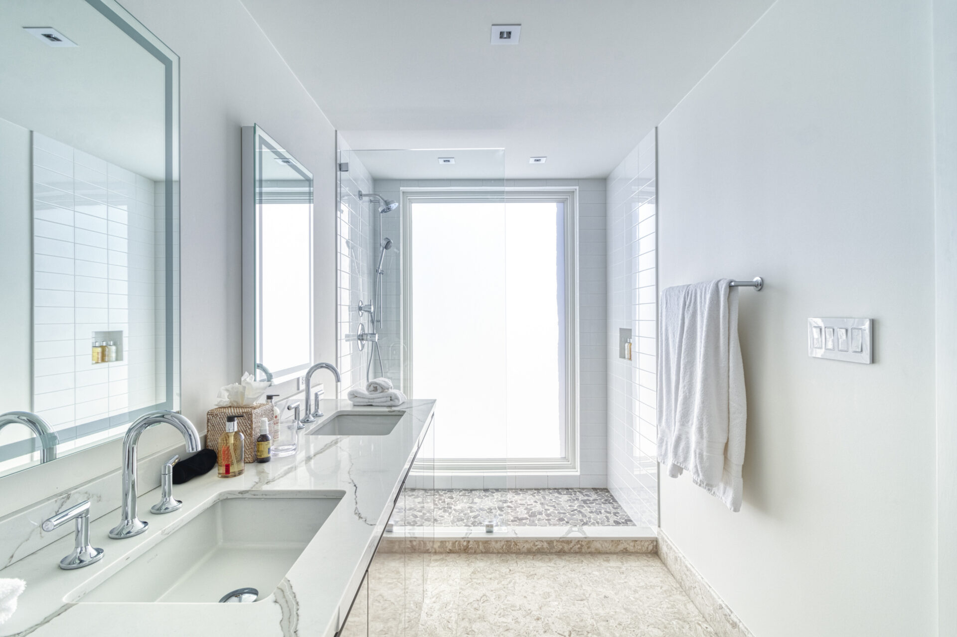 A modern bathroom at Wild Side Villa features a double-sink vanity, large mirrors, and a glass-enclosed shower. The space is adorned with white tile walls and a mix of white and light grey marble countertops. A white towel hangs on a metal bar near the shower while a skylight brightens the room.