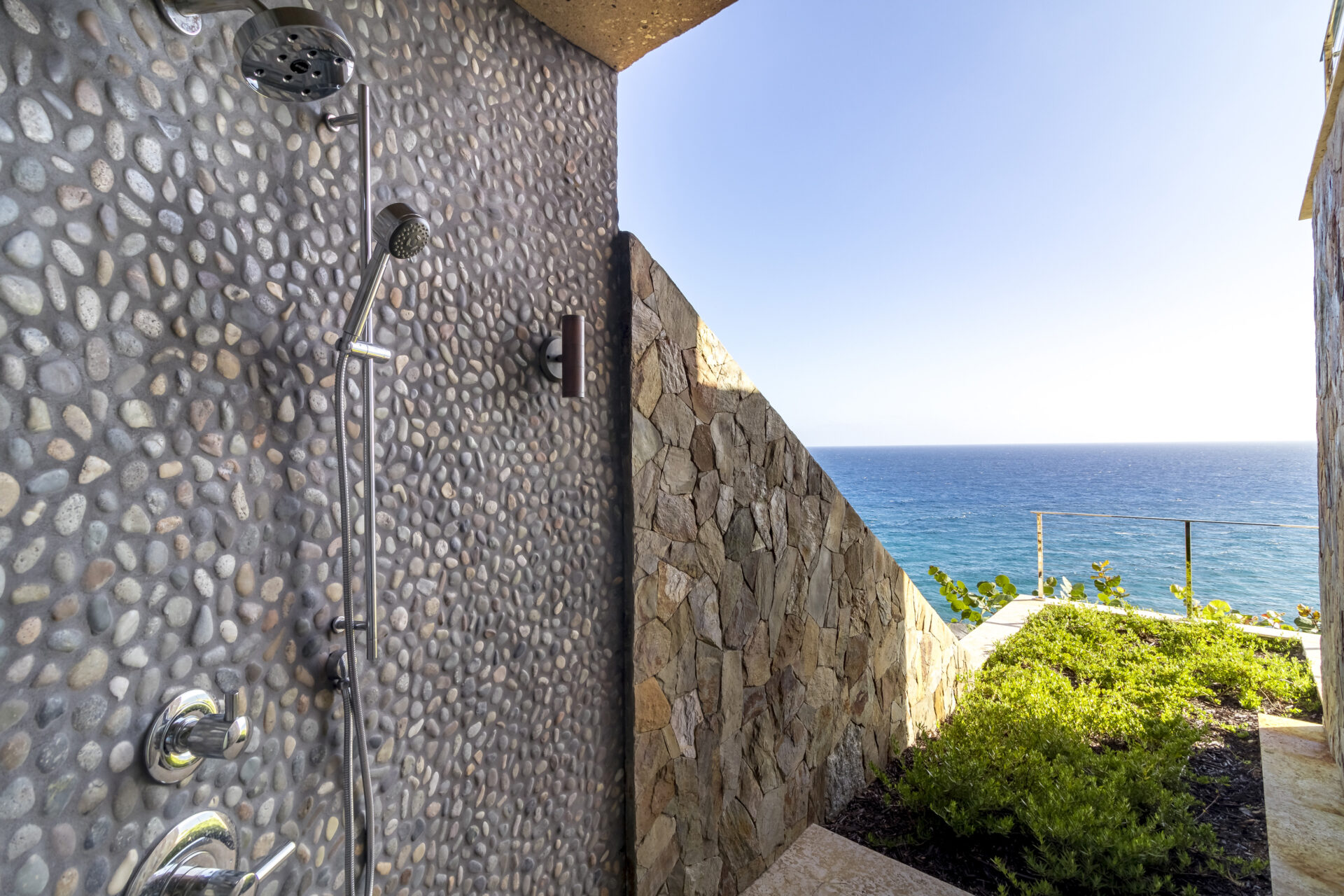 An outdoor shower with a stone wall and pebbled tiles is shown, overlooking a scenic ocean view at Wild Side Villa. The shower hardware is modern with a wall-mounted showerhead and handle. The adjacent area features greenery and a small stone fence near the edge of the cliff.