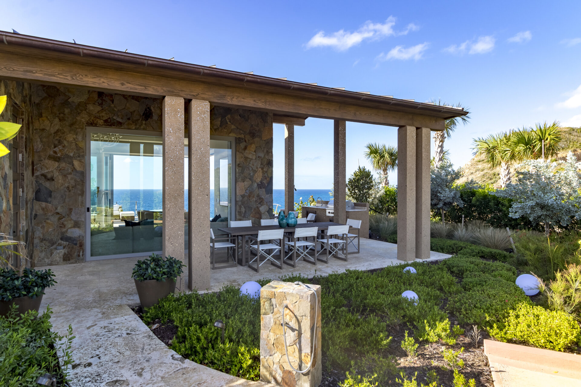 Wild Side Villa is a modern seaside house featuring large stone columns and an outdoor dining area furnished with a white table and chairs. The patio overlooks a garden filled with various plants, offering an unobstructed view of the ocean and blue sky in the background.