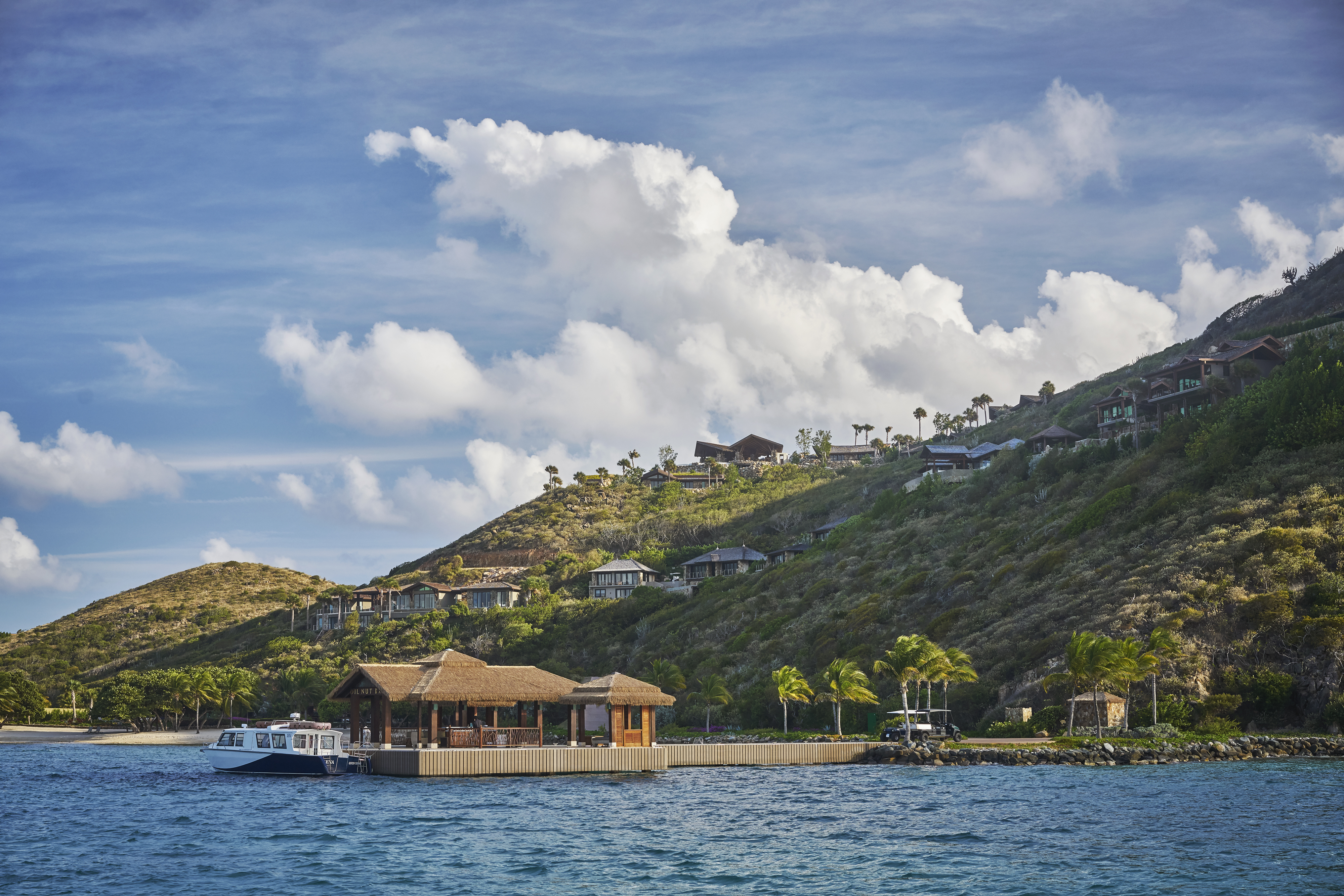 A serene, tropical beach scene featuring a thatched-roof hut on a small dock, with a white boat moored nearby. Green hills with various buildings dotting the landscape rise in the background under a sky filled with fluffy clouds, reminiscent of the best luxury resorts in the BVI.