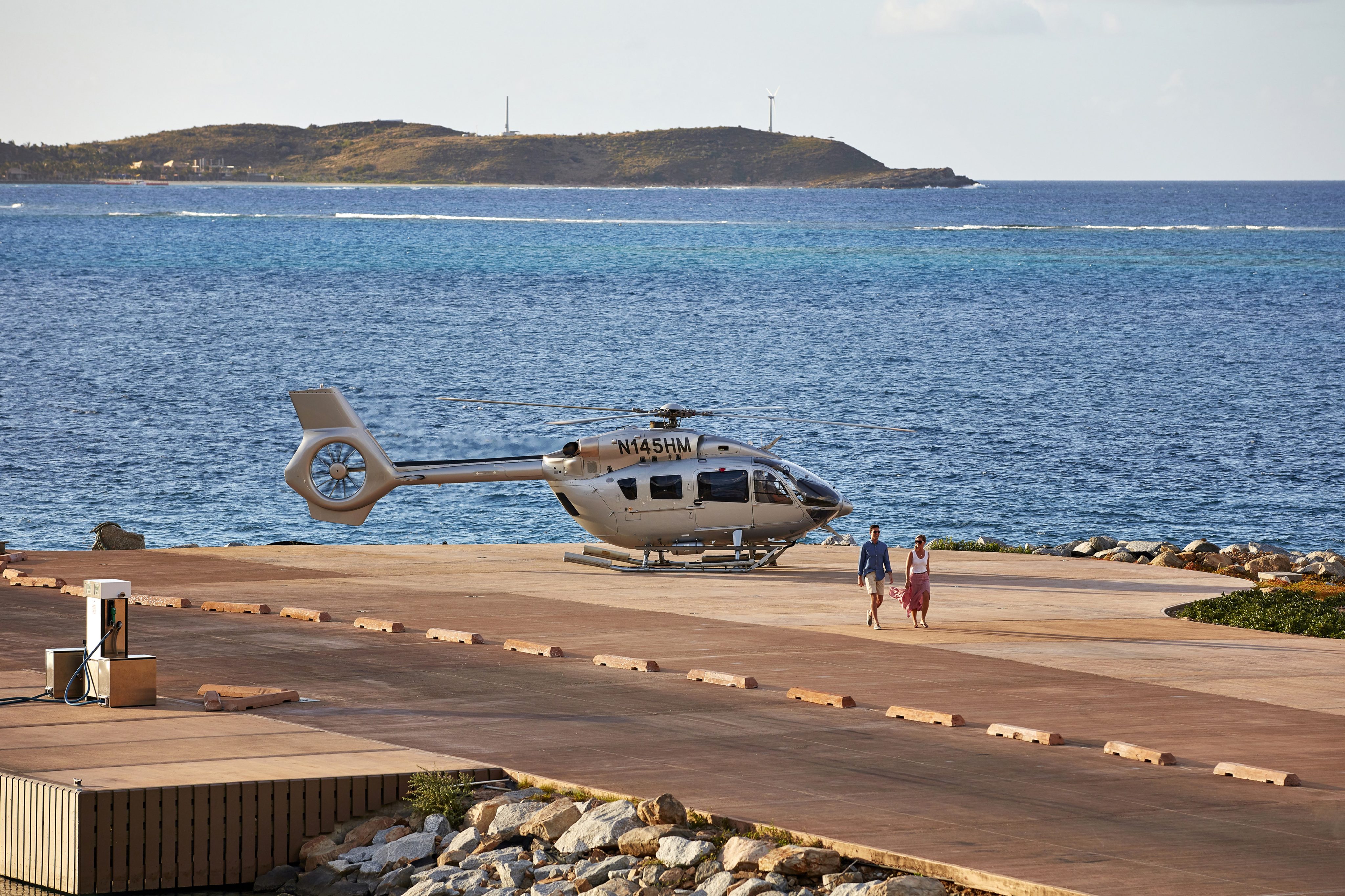 A helicopter is parked on a concrete helipad by the ocean, hinting at the nearby best luxury resorts in the BVI. Two people are walking away from the helicopter. An island is visible in the background. The weather is clear with partly cloudy skies.