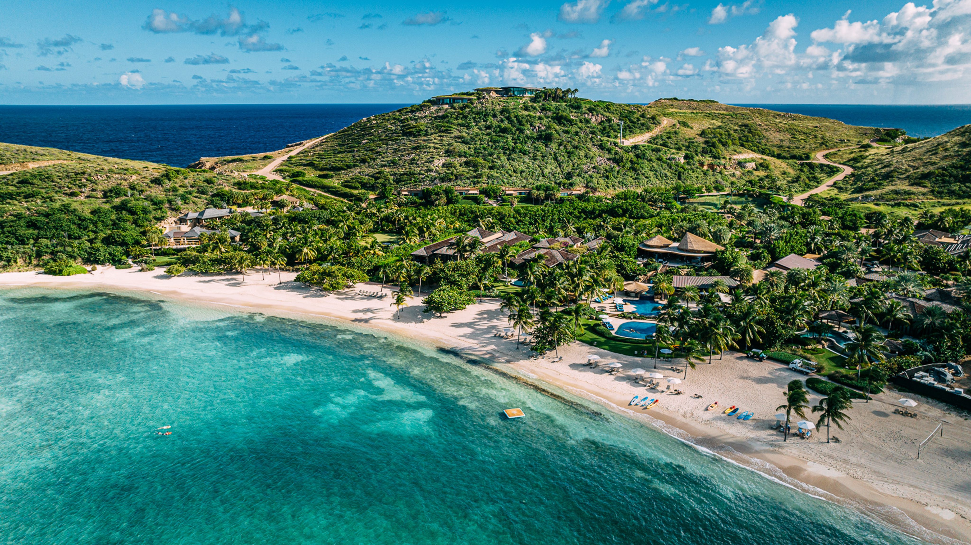 Aerial view of a tropical beach with white sand and turquoise water. Some of the best luxury resorts in the BVI are nestled among lush green hills and palm trees. The ocean extends to the horizon under a blue sky with scattered clouds, where a few people are relaxing on the beach.