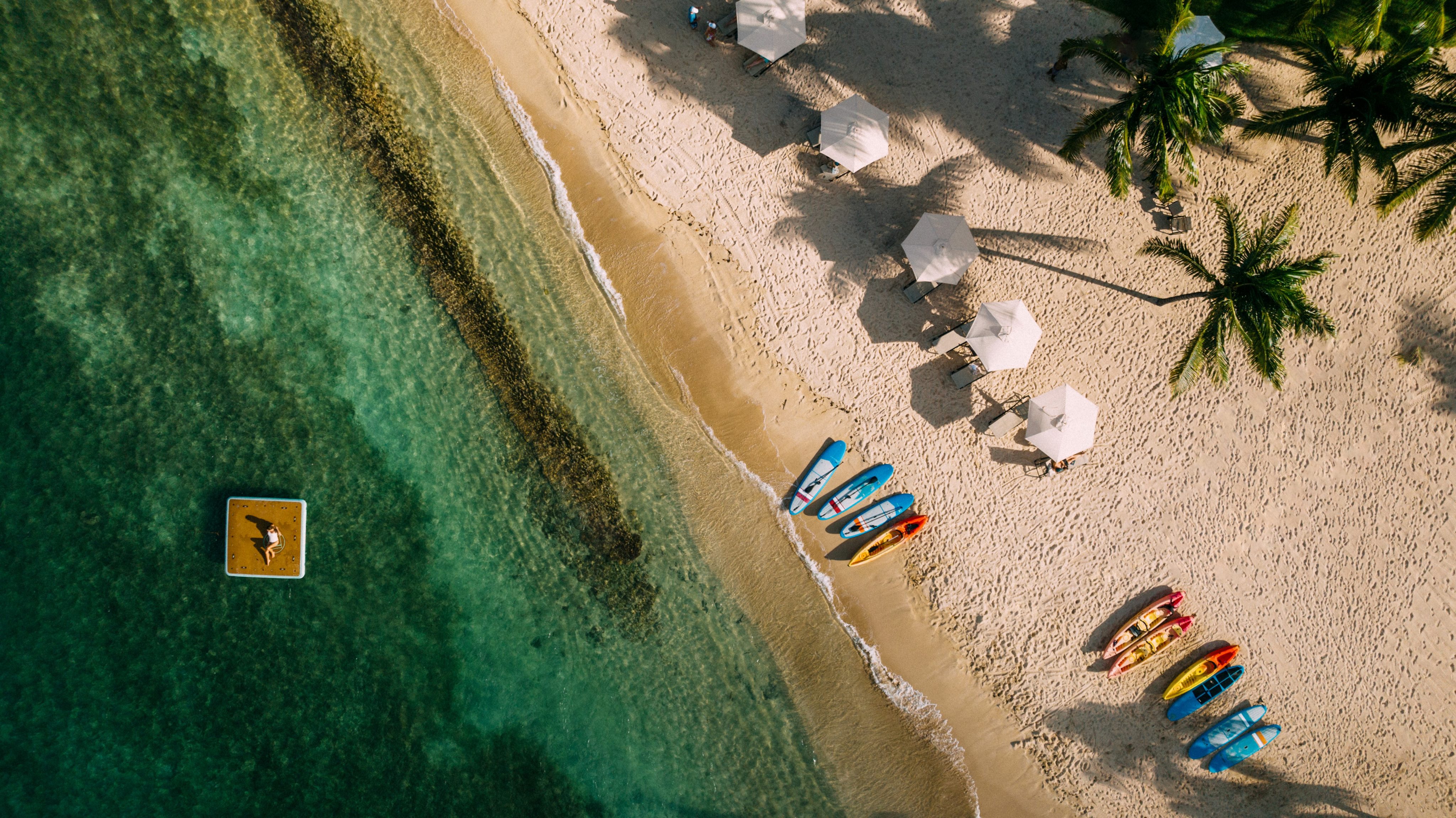 Aerial view of a tropical beach with clear turquoise waters at one of the best luxury resorts in the BVI. Paddleboards and kayaks are lined up on the sandy shore. White umbrellas provide shade to lounge chairs spread along the beach. A person can be seen on a paddleboard floating on the water.