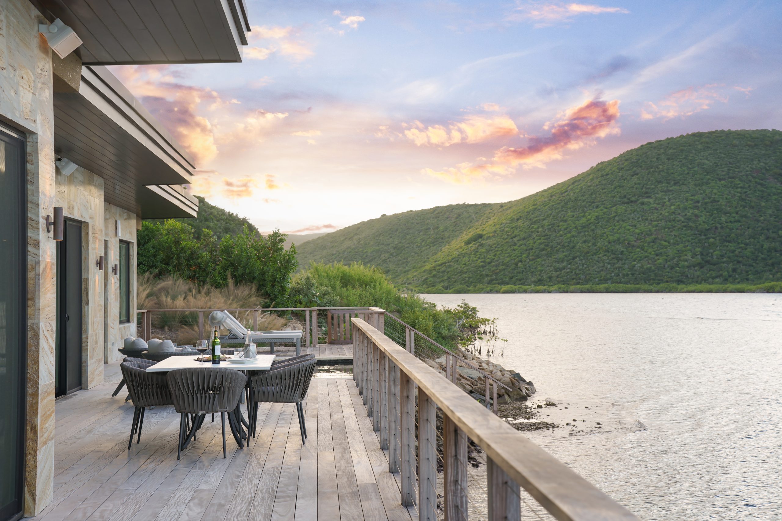 A spacious wooden deck extends from Amanzi Villa, a modern lakeside house at sunset. The deck features a dining table with six chairs and a lounging area with cushioned outdoor furniture. Mountains frame the tranquil lake, and the sky is painted with colorful clouds.