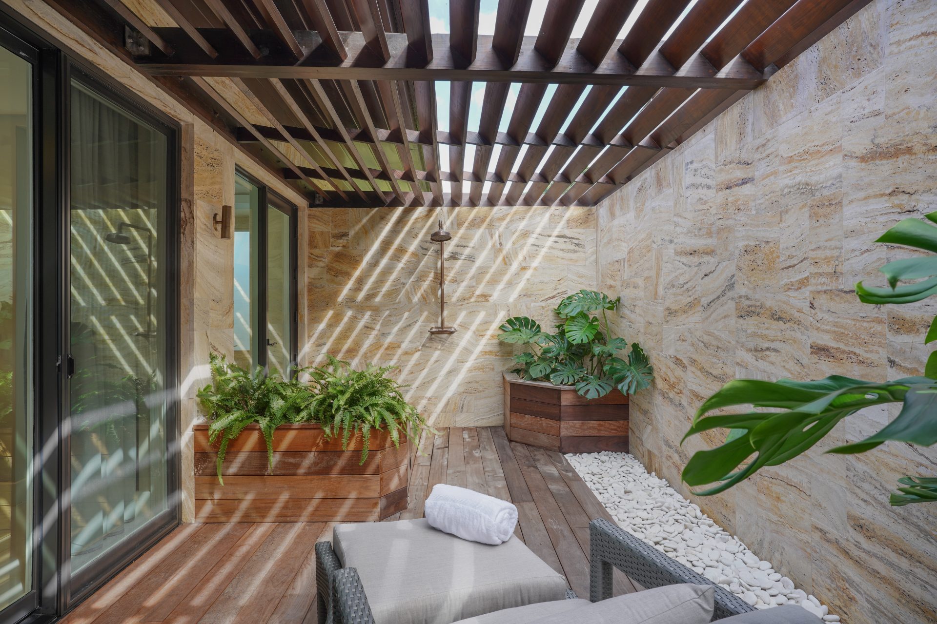 A serene outdoor patio at Amanzi Villa features a wooden pergola casting striped shadows on a stone wall. This luxury accommodation includes a lounge chair with a rolled towel, two wooden planters with lush green plants, and decorative white stones lining the wall. Glass doors open to the interior.