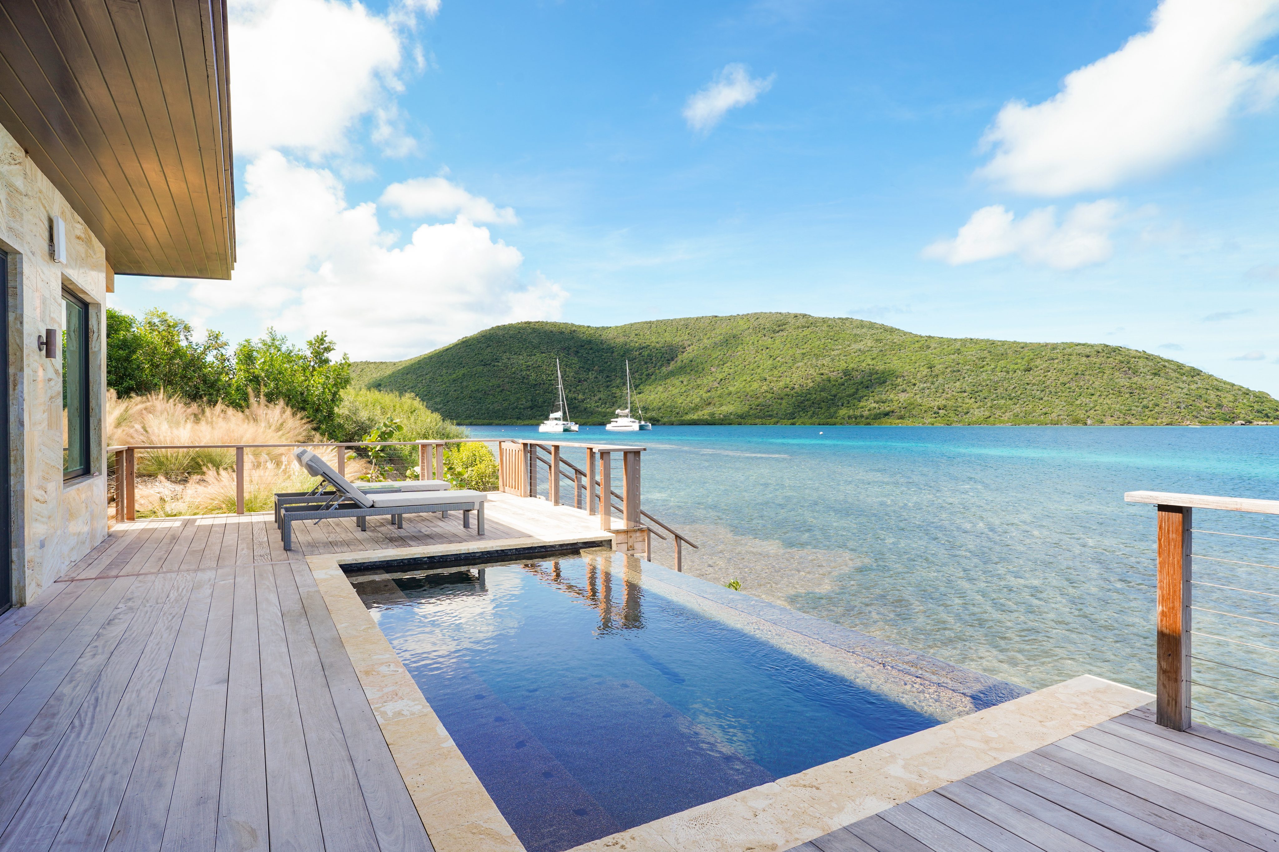 View of a luxurious waterfront patio at Amanzi Villa featuring a small infinity pool, sun loungers, and a wooden deck overlooking a clear blue sea. Two sailboats in the distance float near a lush green hillside under a bright, partly cloudy sky.