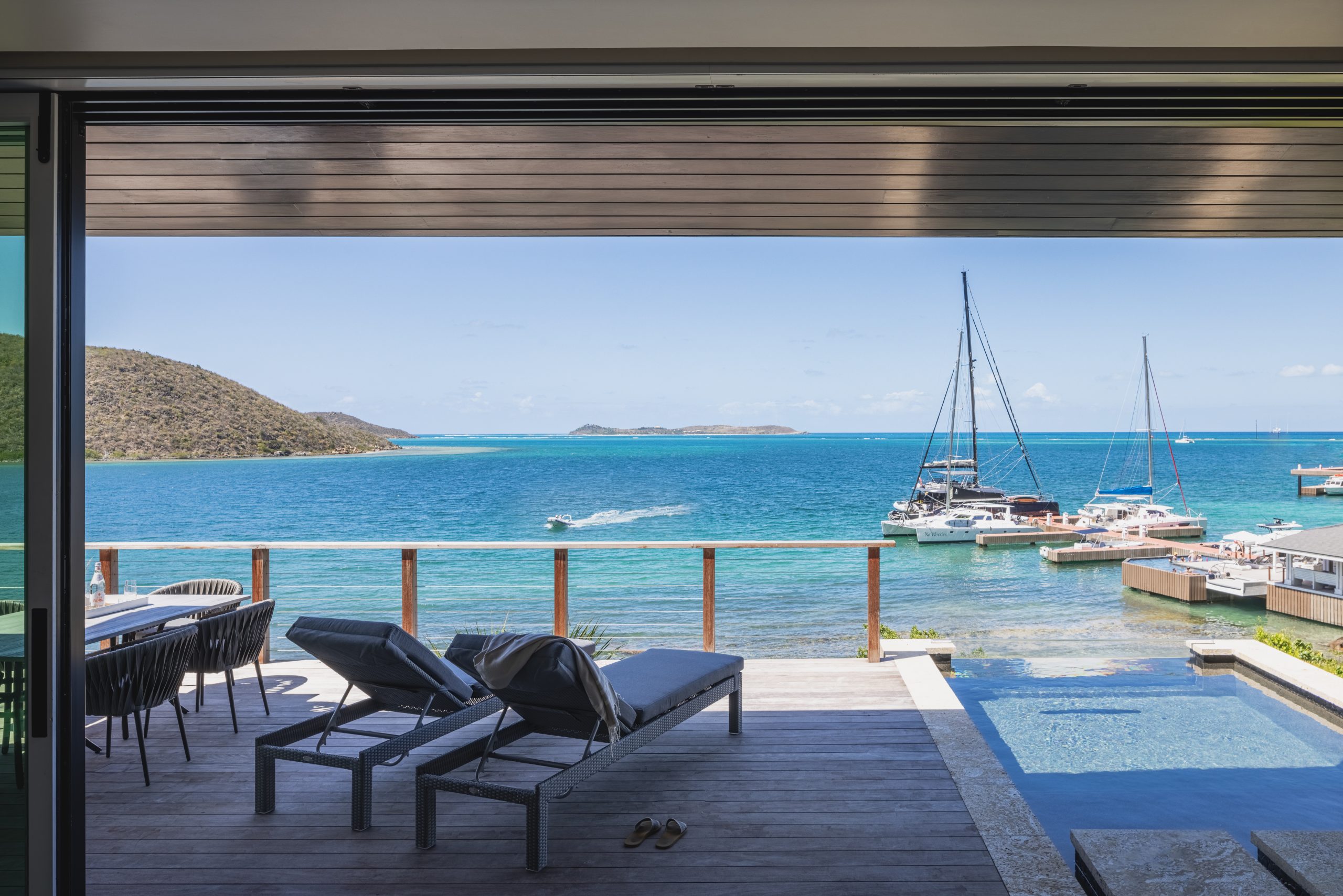 A picturesque view from a modern villa's patio overlooking a calm blue sea with sailboats and yachts docked nearby. The patio features two lounge chairs, a small table with chairs, and an infinity pool. Hills and a distant island are visible across the water, perfect for sipping Amarone as the sun sets.