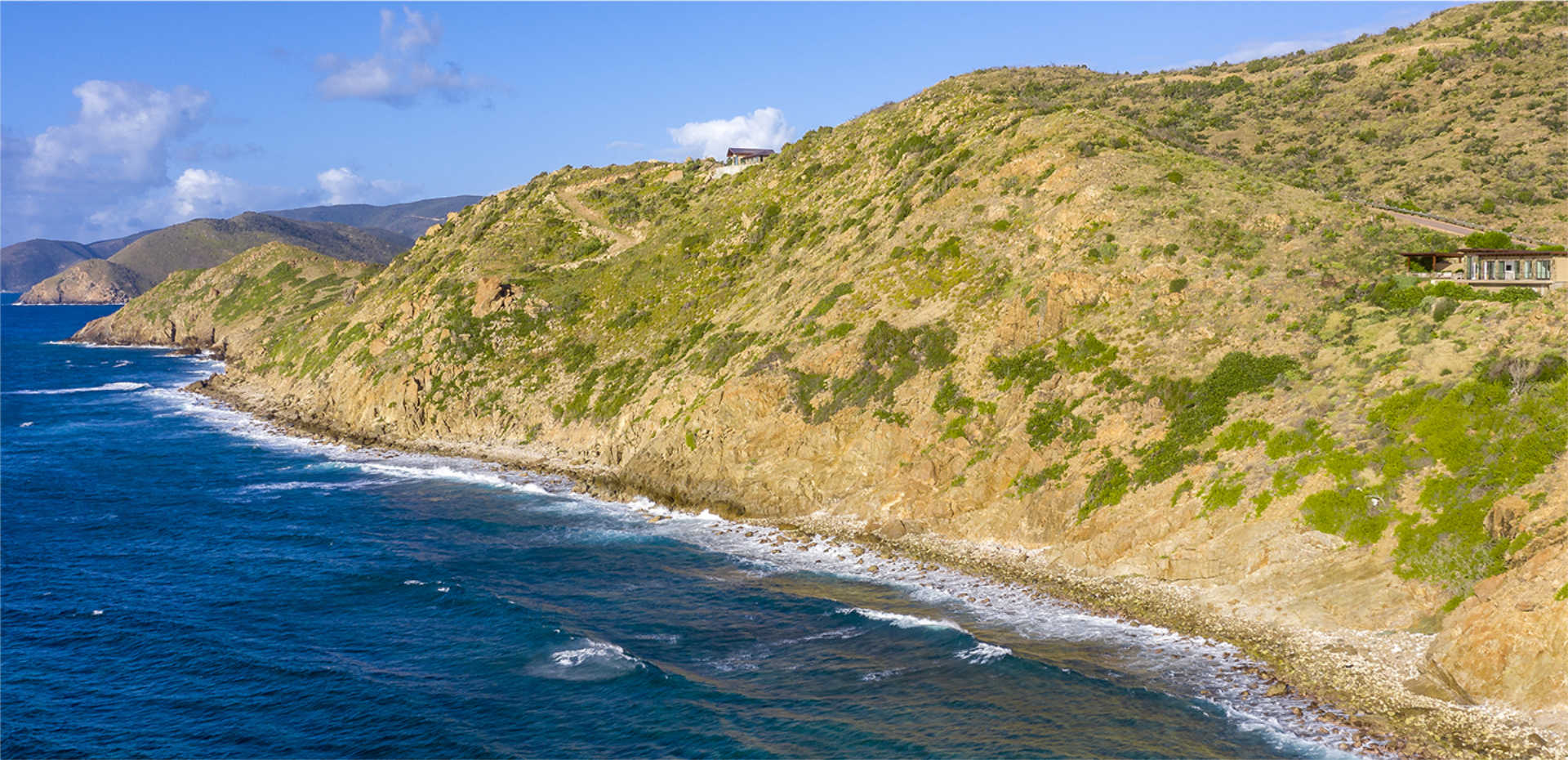 A coastal landscape featuring a rocky shoreline with green hills gently sloping into the blue ocean, where Atlantic Ridge Homesite A12 sits nestled. Small waves break against the shore, and the sky is partly cloudy, casting soft light on the scene, creating a peaceful and scenic view.