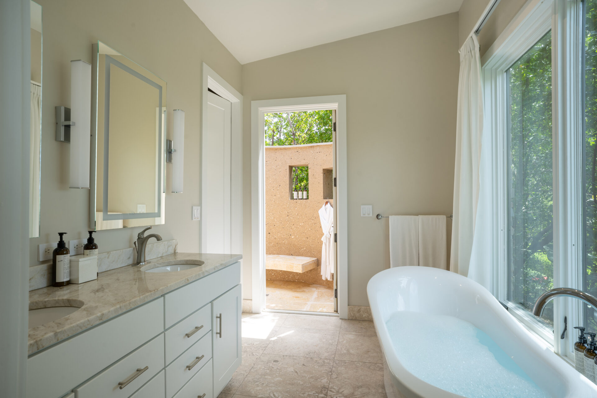 A luxurious bathroom suite with beige walls and marble countertops. It includes a double sink vanity with two mirrors, white cabinets, and a freestanding bathtub partially filled with water. Large bay windows bring in natural light. The open door reveals a patio area outside.