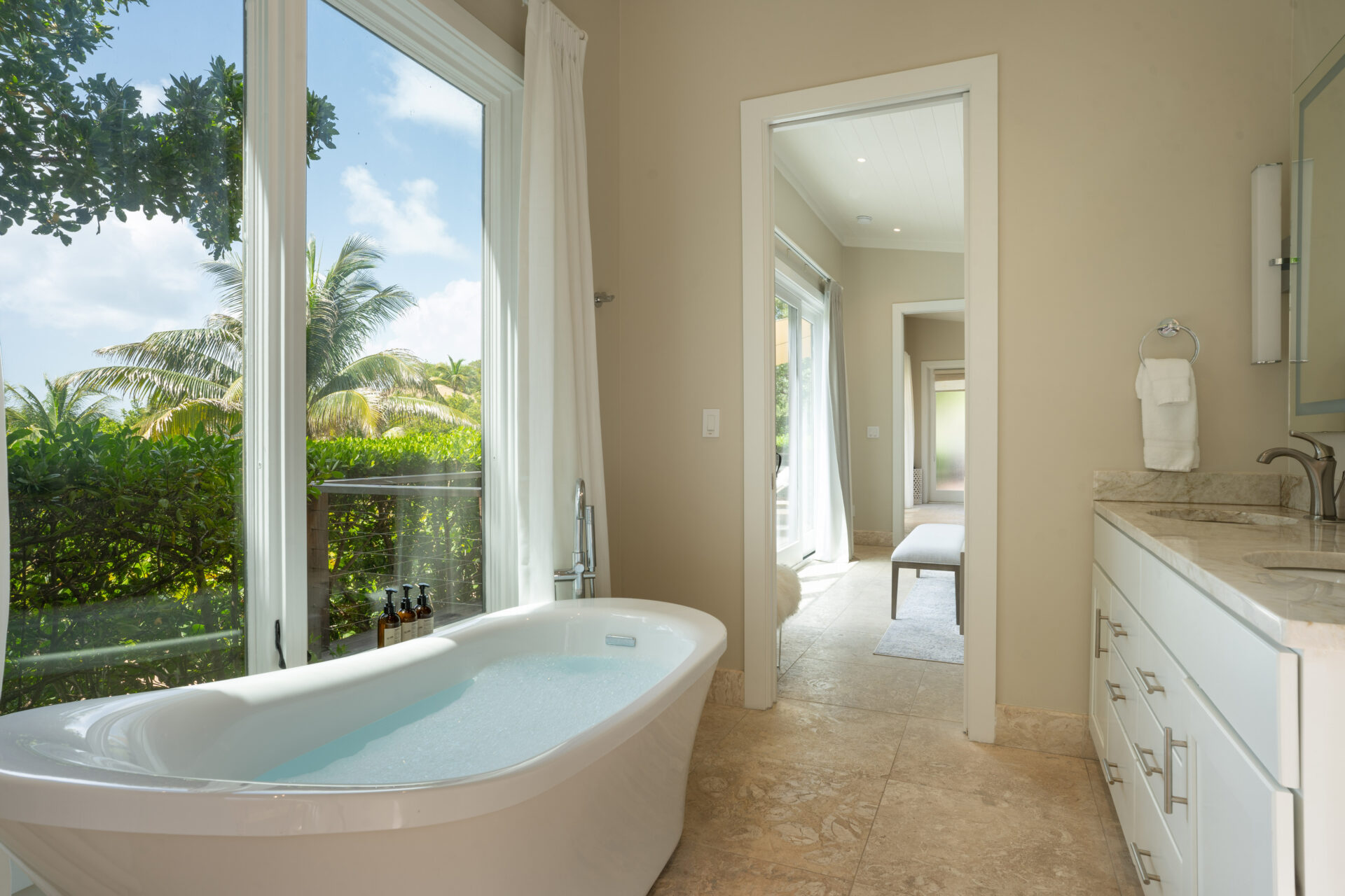 A serene bathroom in the Bay Suites features a freestanding bathtub next to large windows offering a lush garden view. The room includes a long, white vanity with a marble countertop, a mirror, and a doorway leading to a sunlit hallway. The decor is minimalist and fresh.