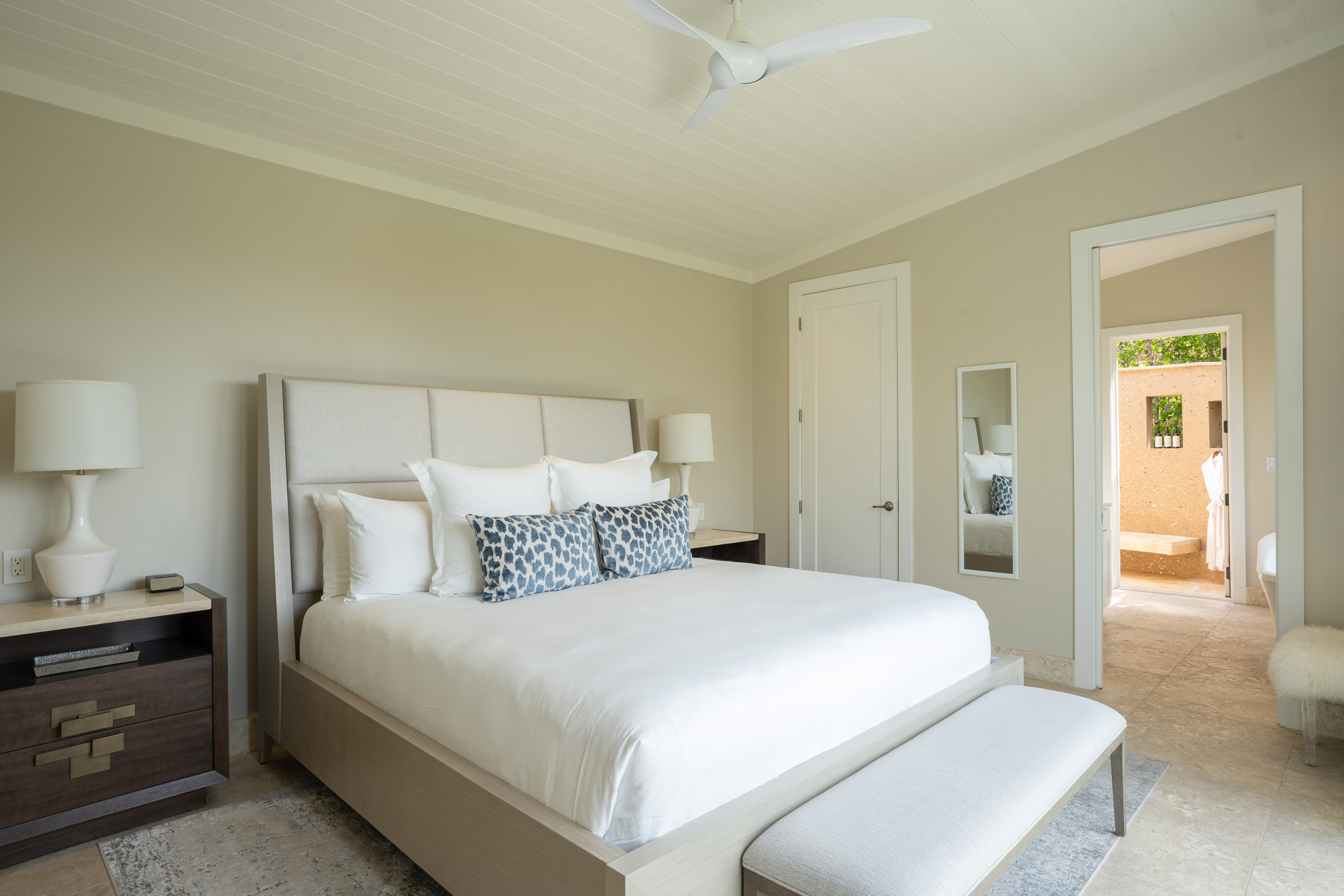 A serene bedroom with a large bed adorned with white linens and patterned blue pillows. Two nightstands with lamps flank the bed, and a beige bench is at the foot. A door leads to a bright, open bathroom in one of our luxurious Bay Suites, and a full-length mirror stands against the wall. A ceiling fan is above.