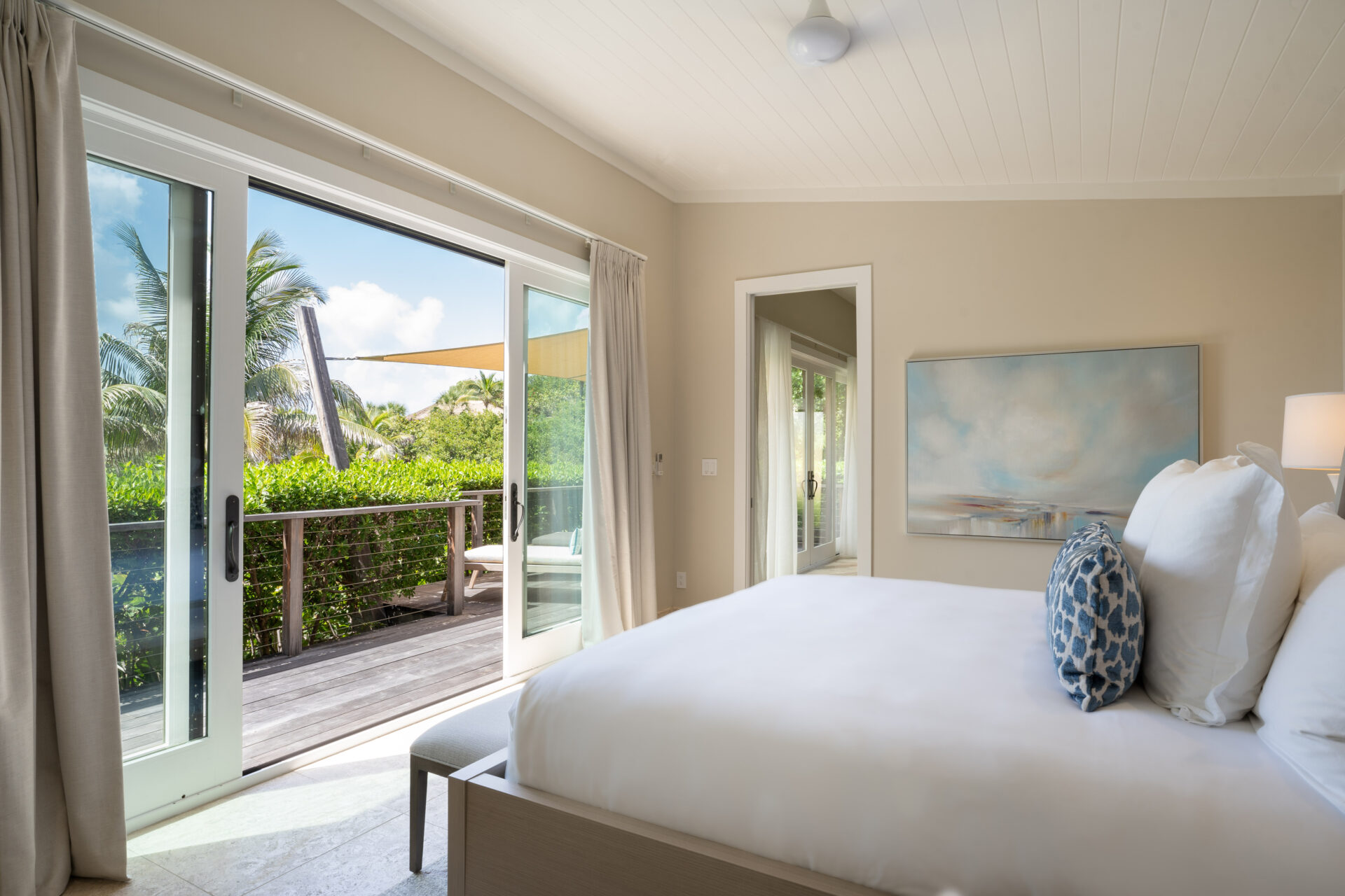 A bright, airy Bay Suites bedroom featuring a white bed with blue accent pillows, light beige walls, and a painting. French doors open to a wooden deck with greenery and palm trees, letting in ample natural light. A white ceiling fan and soft beige curtains complete the serene look.