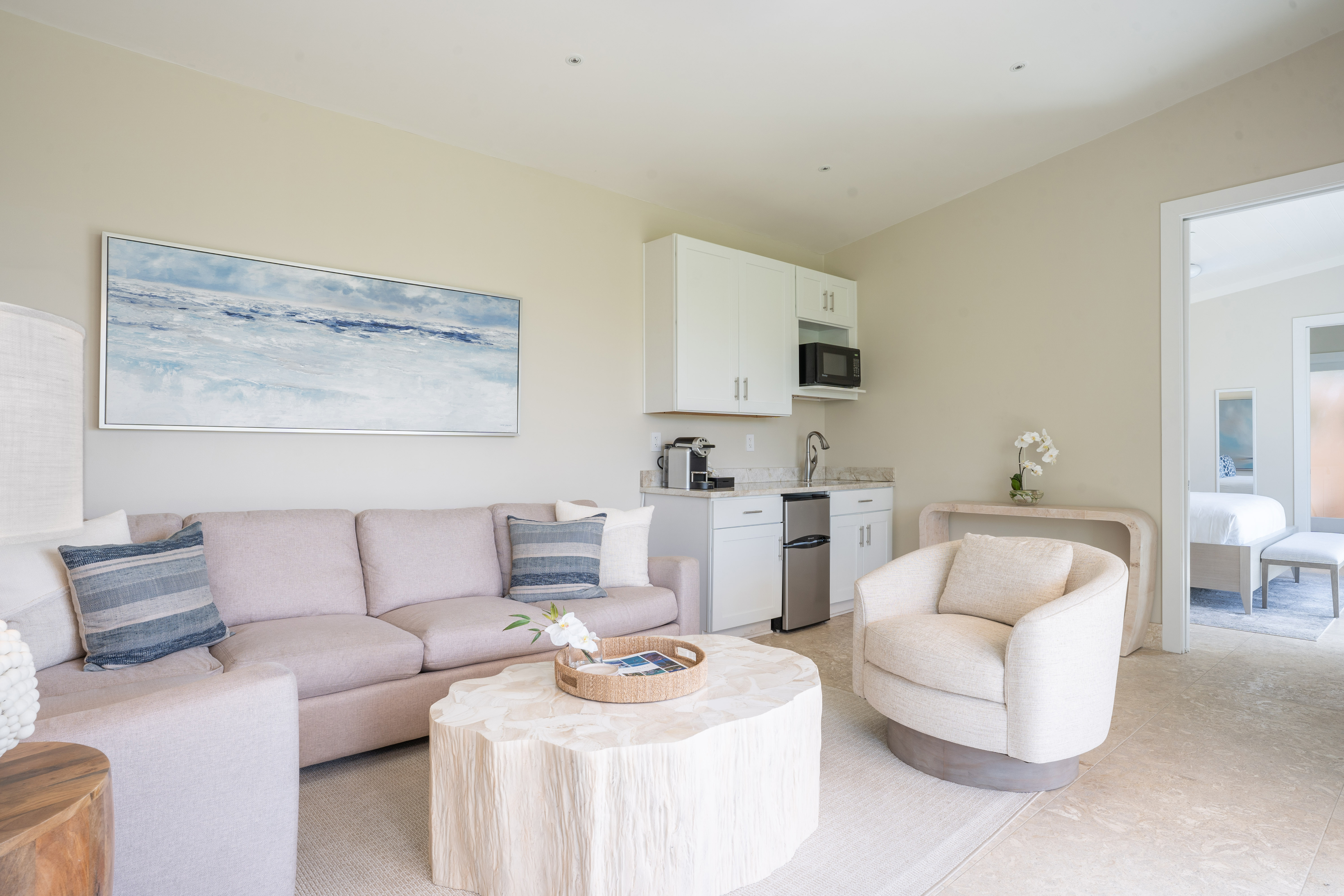 A modern living room at Bay Suites features a beige sofa, blue and gray pillows, a round white coffee table, a white armchair, and wall art showcasing an abstract seascape. The kitchenette is equipped with white cabinets, a microwave, and a coffee maker. A doorway leads to the bedroom.