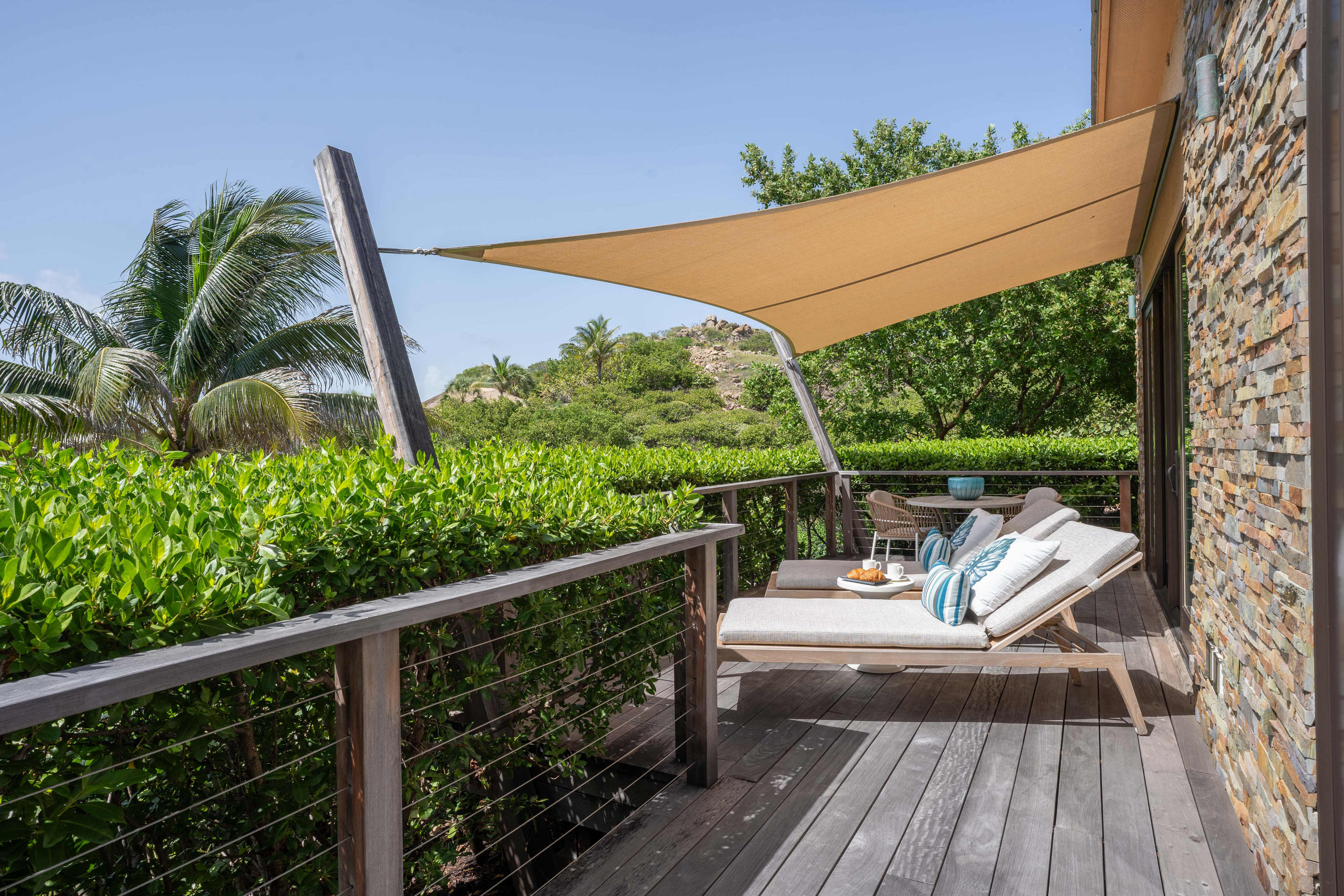 A sunshade covers two lounge chairs on a wooden deck, overlooking lush greenery and palm trees at Bay Suites. A small table holds a drink and a snack tray between the chairs. The side of a stone-clad building is visible on the right. The sky is clear and blue.