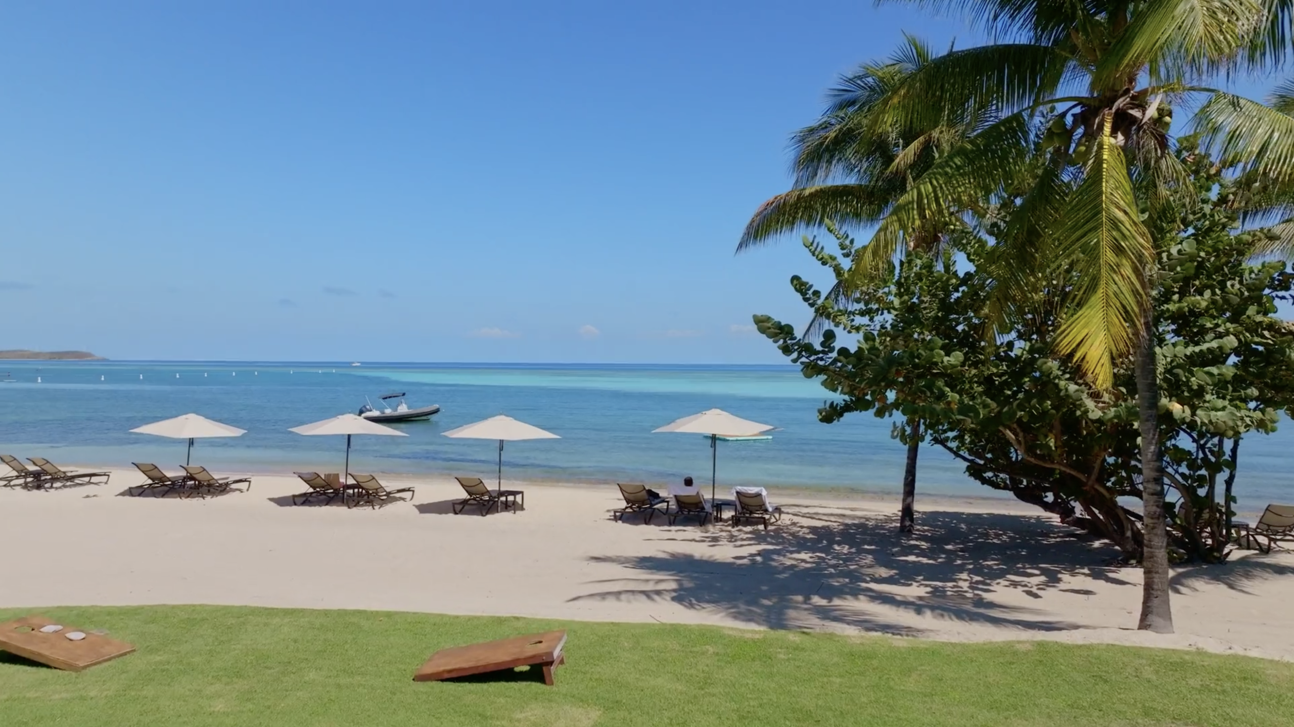 A tranquil beach scene at the bay with clear blue water, a few boats in the distance, and several lounge chairs under white umbrellas on the sandy shore. A large palm tree provides additional shade, and the sky is clear with minimal clouds. Lush green grass is in the foreground.