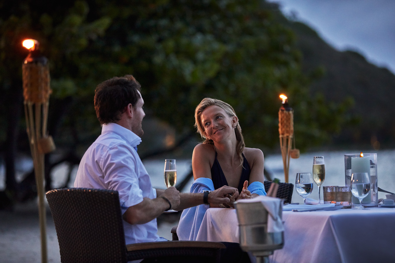 A couple sits at a candlelit outdoor table on a beach club at dusk, holding hands and looking at each other lovingly. They are surrounded by torches, with glasses of champagne and dinnerware on the table. Trees and the ocean are visible in the background.