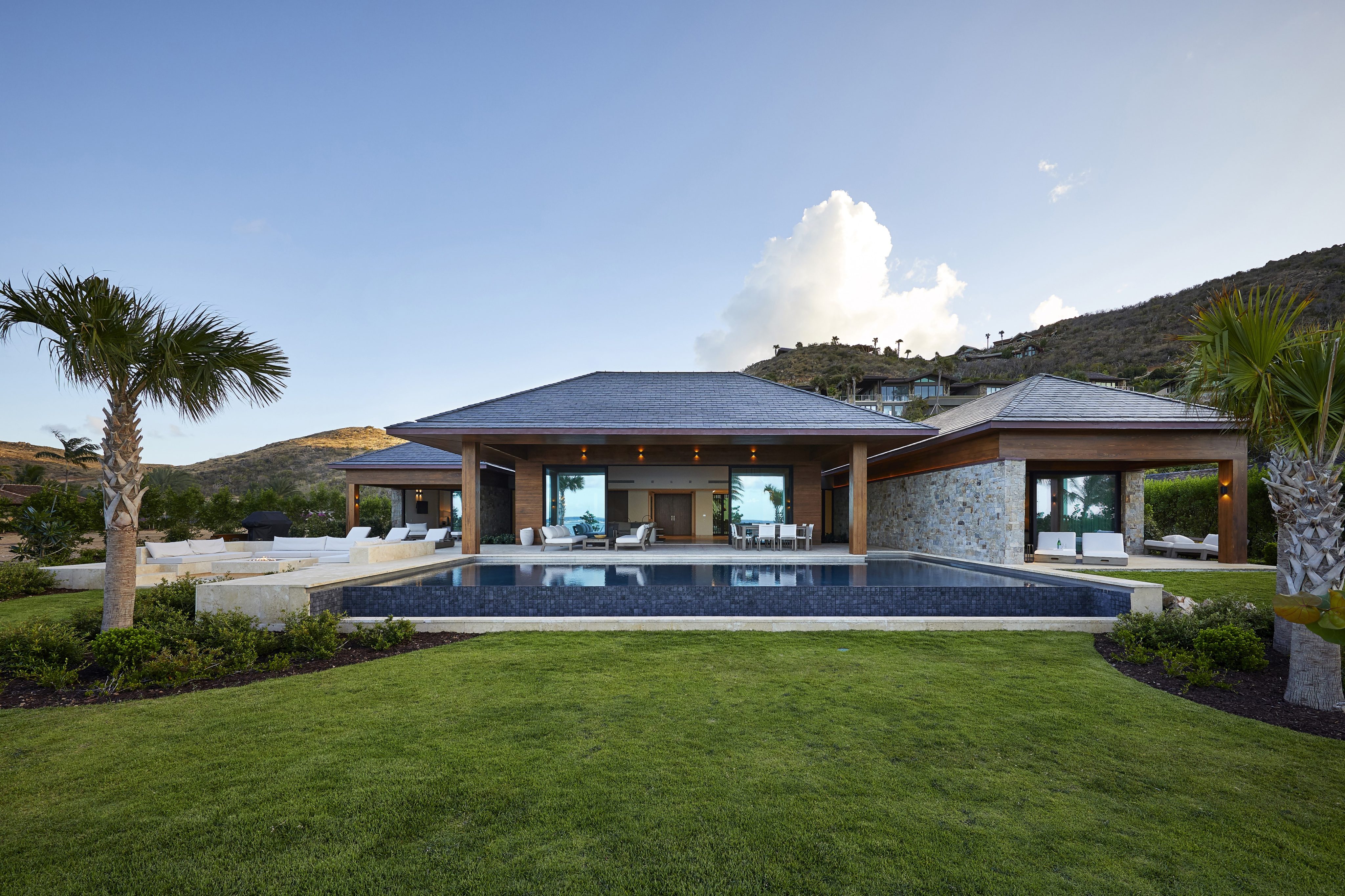 A modern beach house with large glass windows and wooden accents, featuring a spacious lawn, a rectangular outdoor pool, and a patio with outdoor furniture. The beachfront property is surrounded by greenery, with hills in the background under a clear sky.