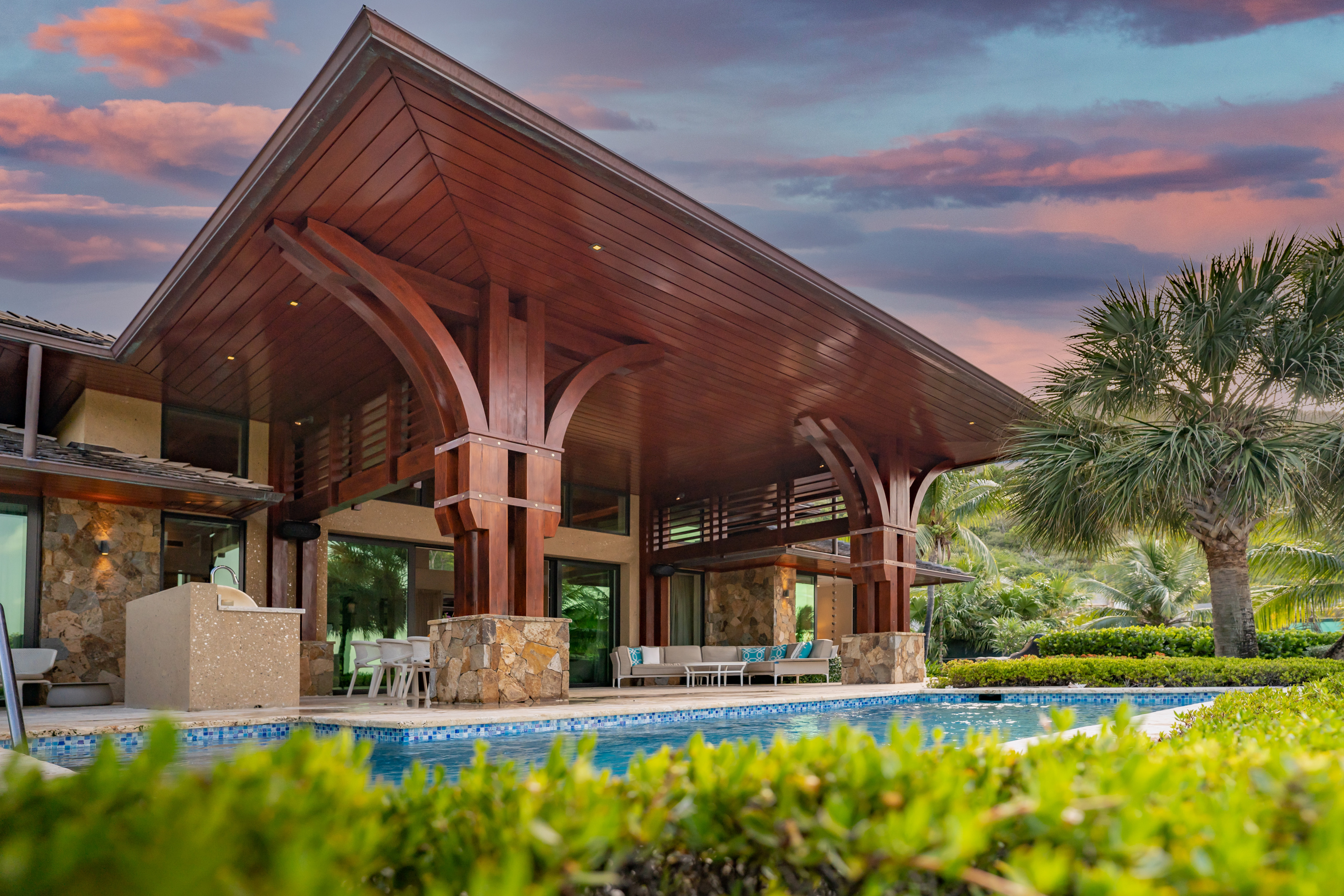 A luxurious modern villa with wooden accents and large glass windows, featuring a covered outdoor seating area with sofas and a dining table. In the foreground, there's a clear blue swimming pool surrounded by well-manicured hedges and lush greenery. The sky is painted with warm hues of pink and orange as the sun sets.